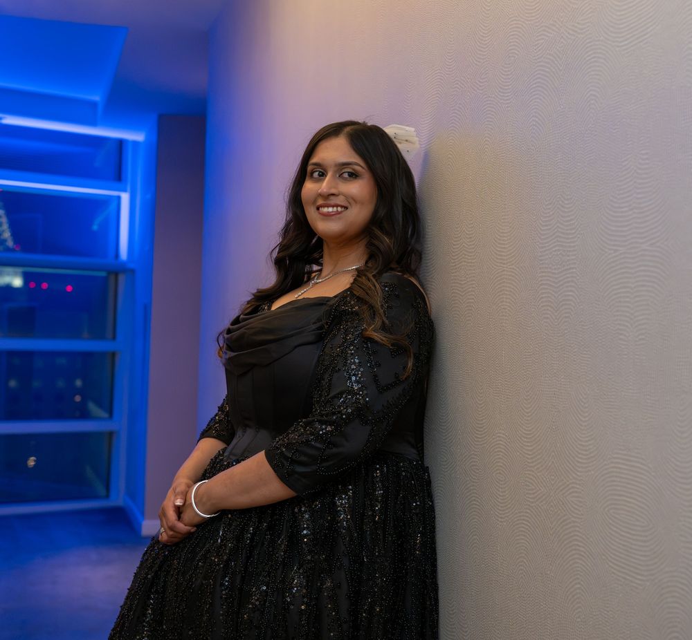 A young woman in a black sequin dress leans against a white wall, smiling, with blue ambient lighting behind her.