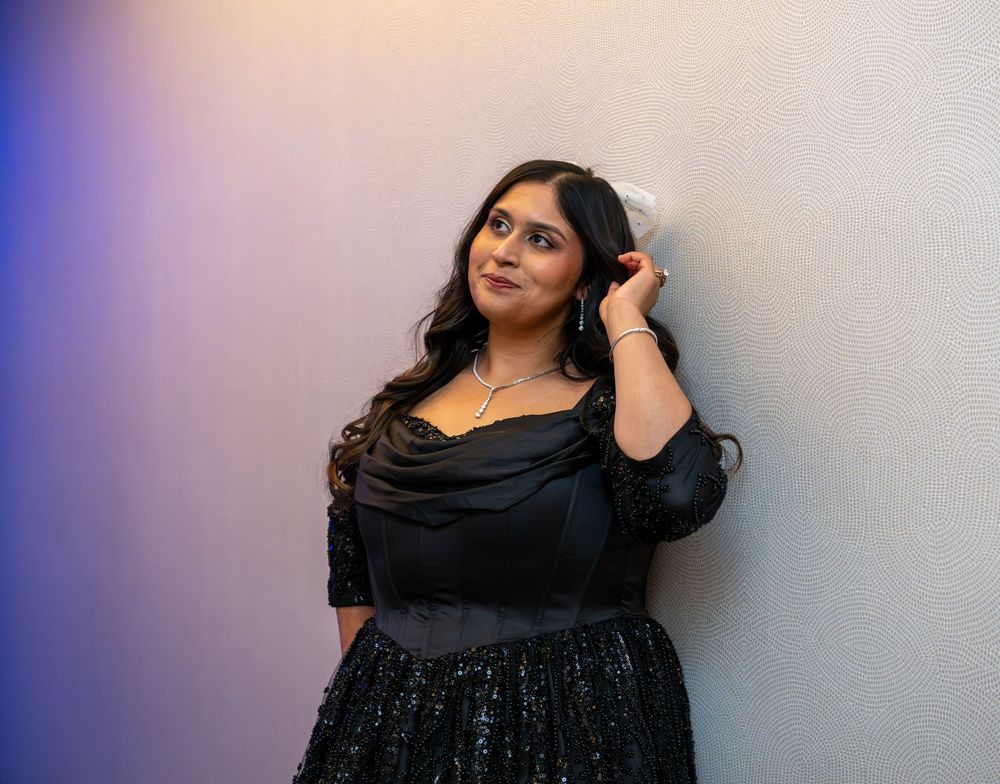 Woman in black off-shoulder sequin dress posing elegantly against a light wall with colorful ambient lighting.
