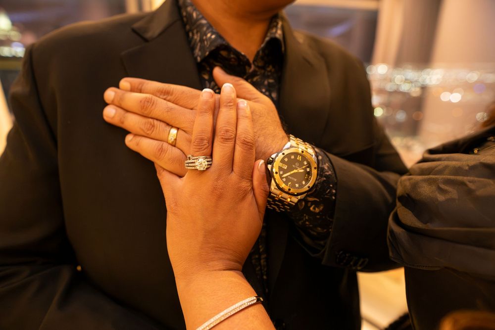 Close-up of two hands clasped together, one wearing a gold ring and bracelet, the other a gold luxury watch.