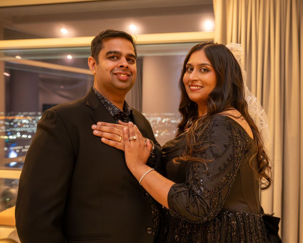 Engaged couple posing together in elegant black attire with city lights visible behind them.