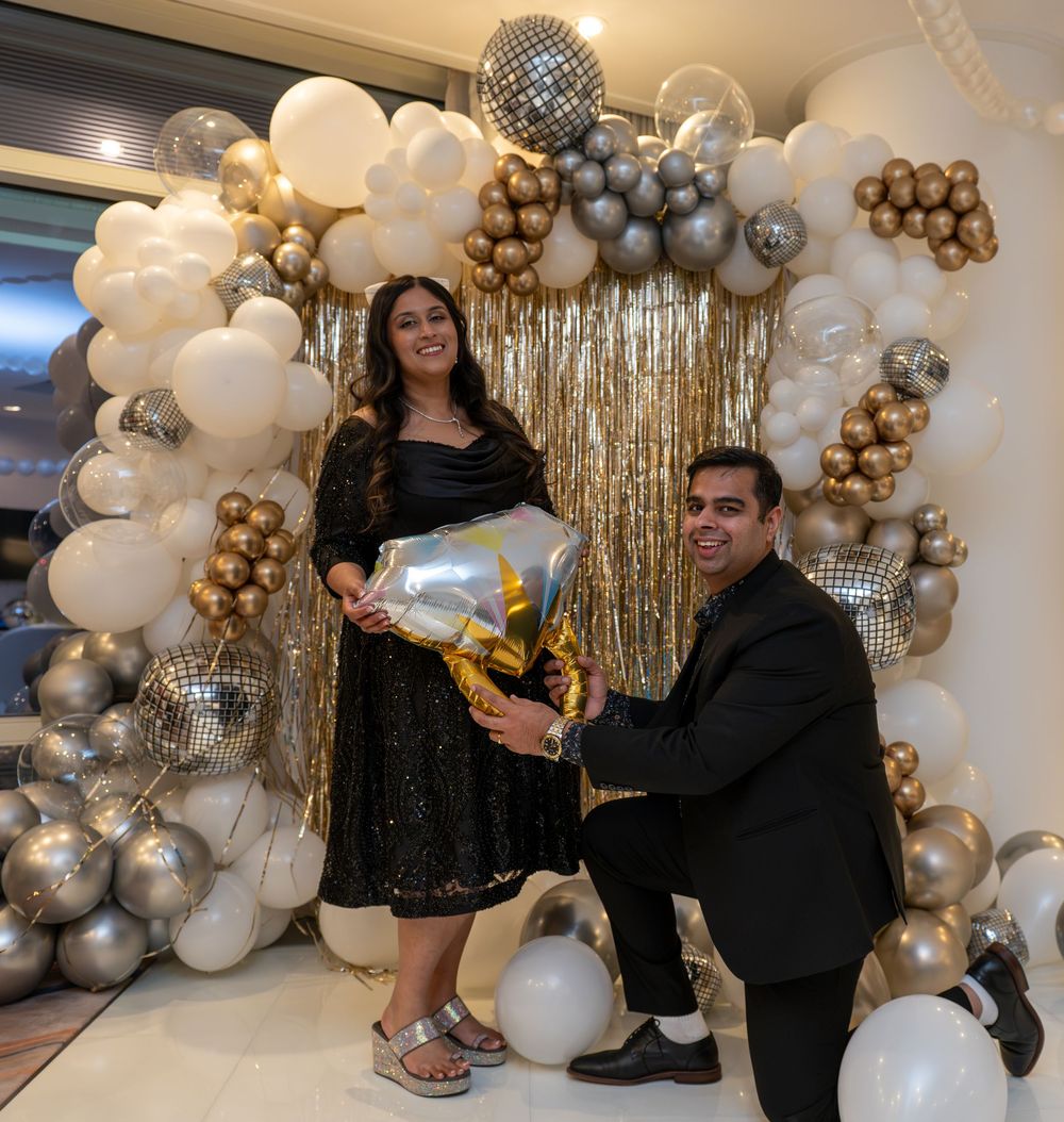 Man kneeling to propose to woman in black dress, gold and silver balloon arch backdrop, celebration decor.