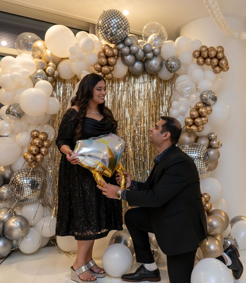 Couple celebrating with balloons and gold decorations, man kneeling presenting a gift to a smiling woman in a black dress.