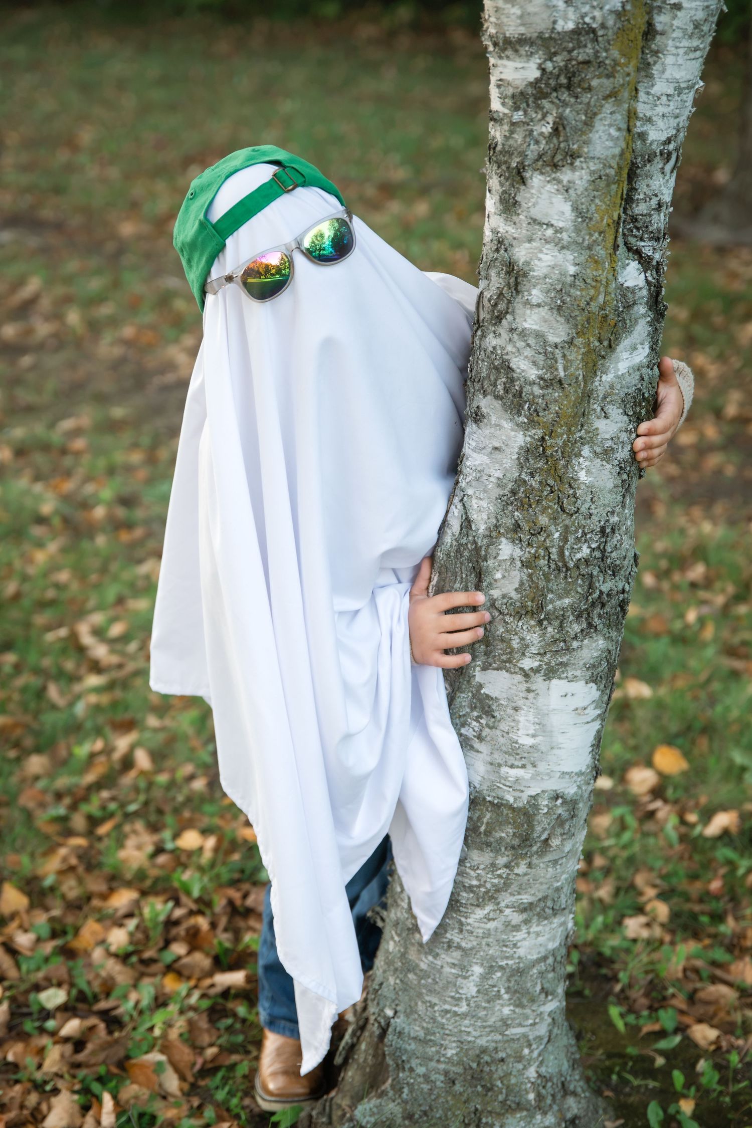 Child in a white sheet ghost costume with green cap and mirrored sunglasses peeking around a birch tree on a leaf-covered lawn.