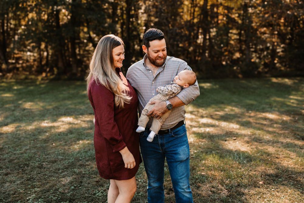 Smiling couple in a sunny wooded park holding their newborn baby during a family photo session in Knoxville, TN.