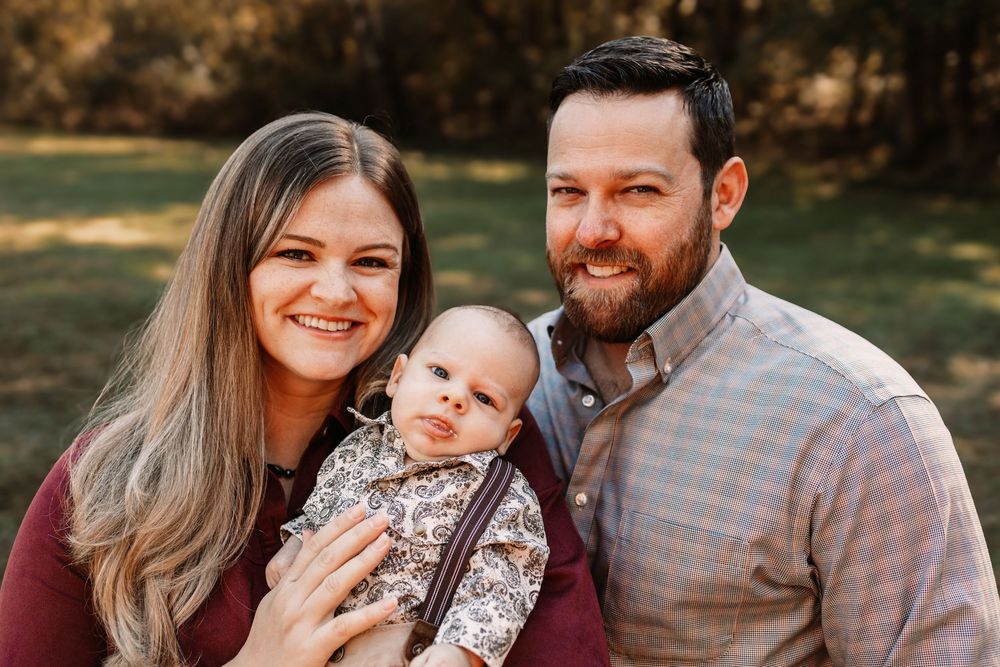 Smiling woman and bearded man in plaid shirt pose outdoors with their baby during a family photo session in Knoxville, TN.