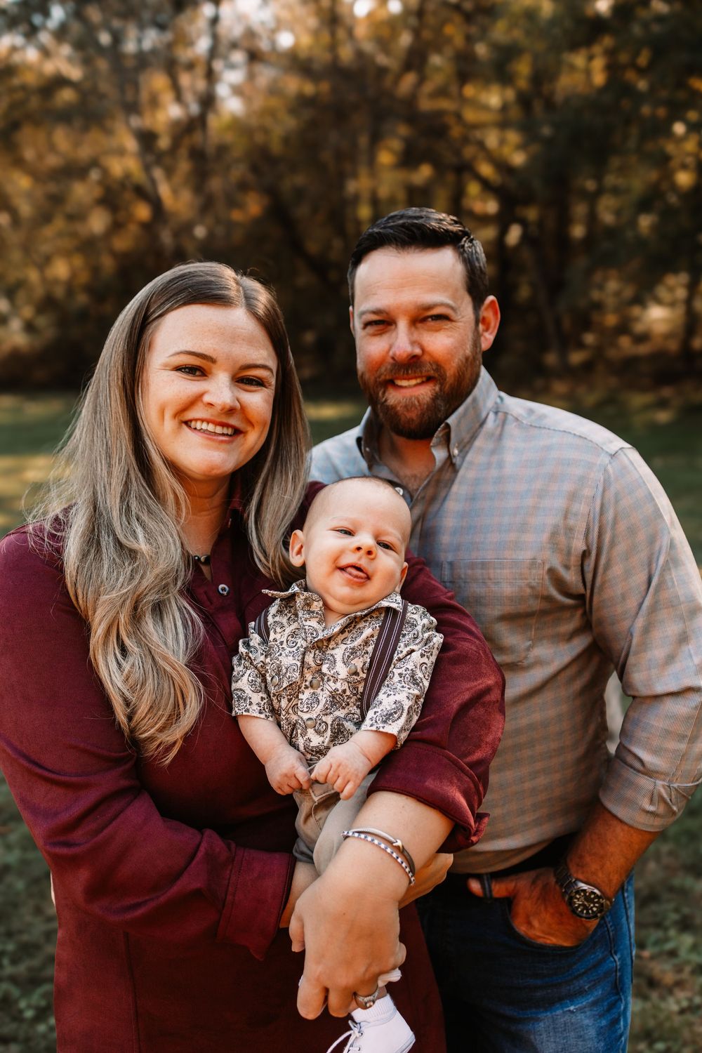Smiling couple in burgundy dress and gray button-up shirt hold their happy baby during a family photo session in Knoxville, TN.