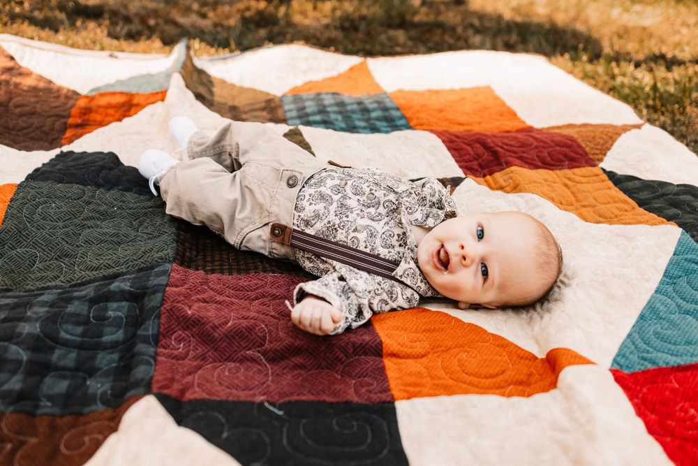 Smiling baby in a patterned outfit and khaki pants lies on a colorful patchwork quilt during a family photo session in Knoxville, TN.