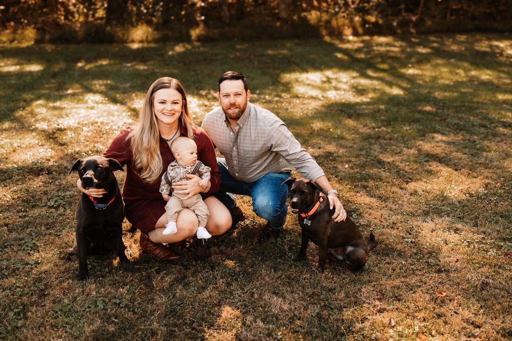 Smiling couple sitting on a sunlit grassy lawn holding their baby during a family photo session in Knoxville, TN.