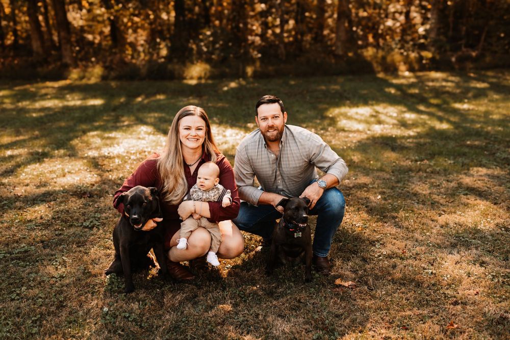 A couple sits on a sunlit grassy lawn with their baby and two black dogs during a family photo session in Knoxville, TN.