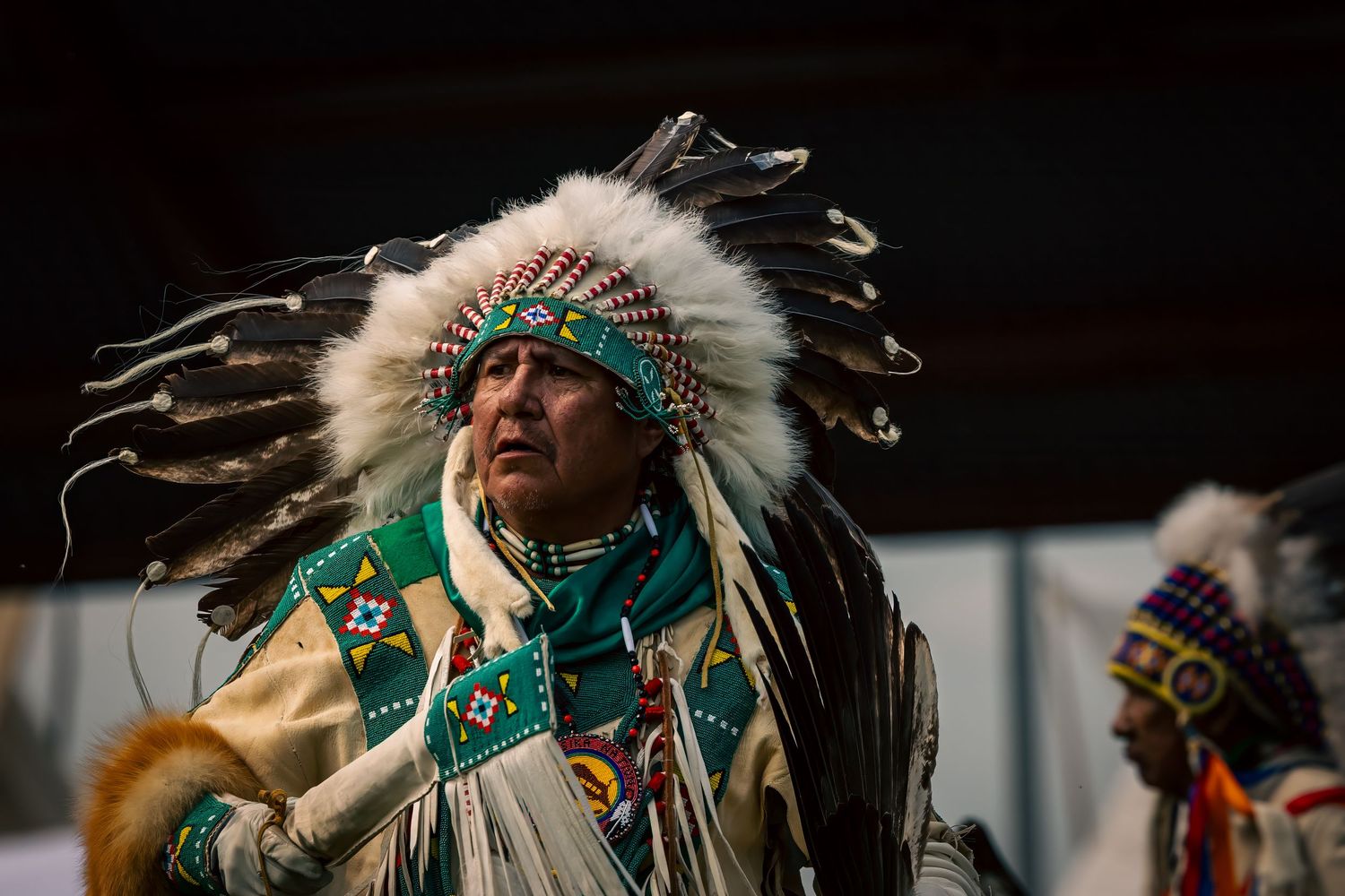 Native American dancer in a feathered headdress and beaded regalia performs at a powwow, with another dancer in colorful attire nearby.