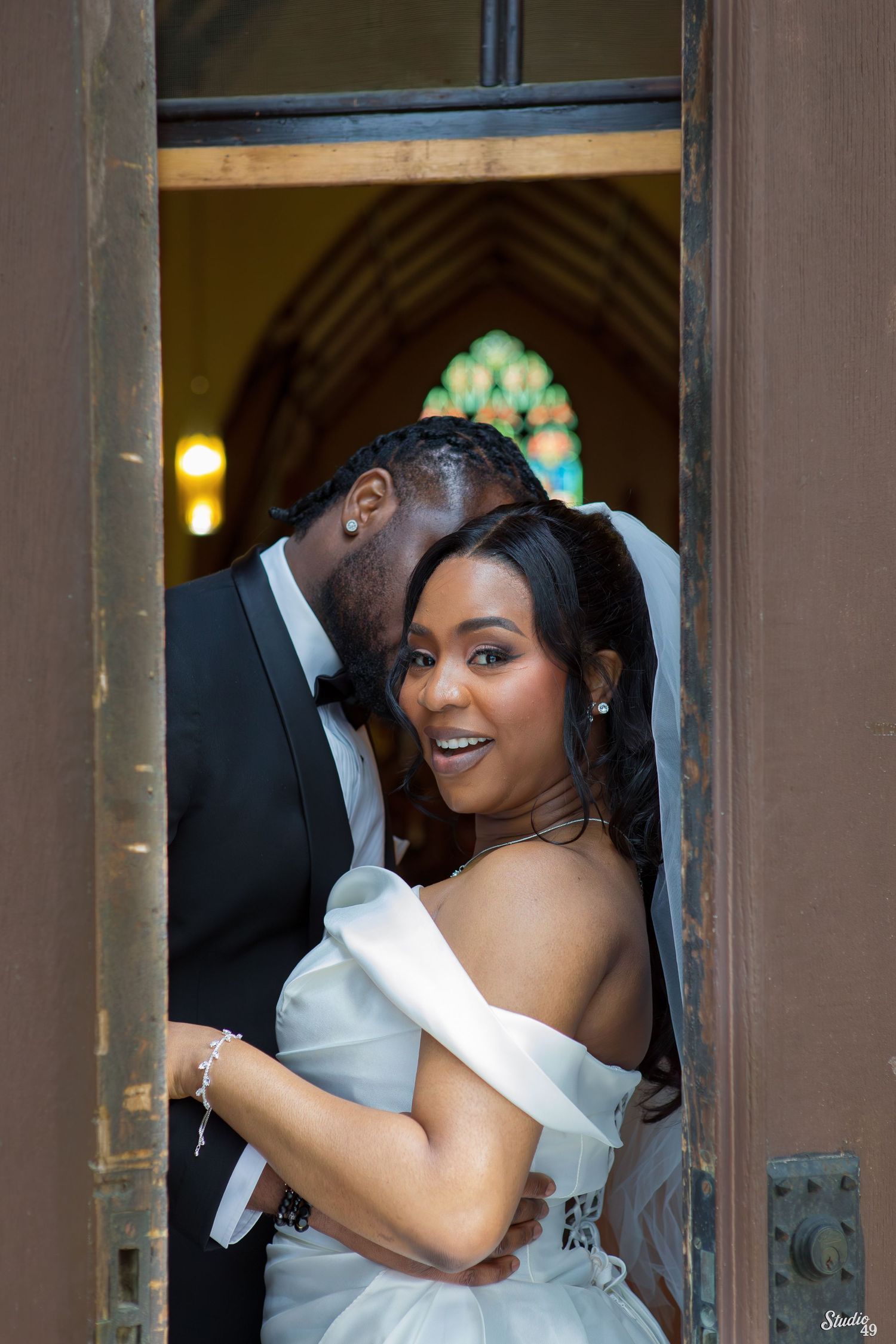 Bride in an off-shoulder white gown smiles as her groom in a black tuxedo embraces her in a church doorway with stained glass behind.