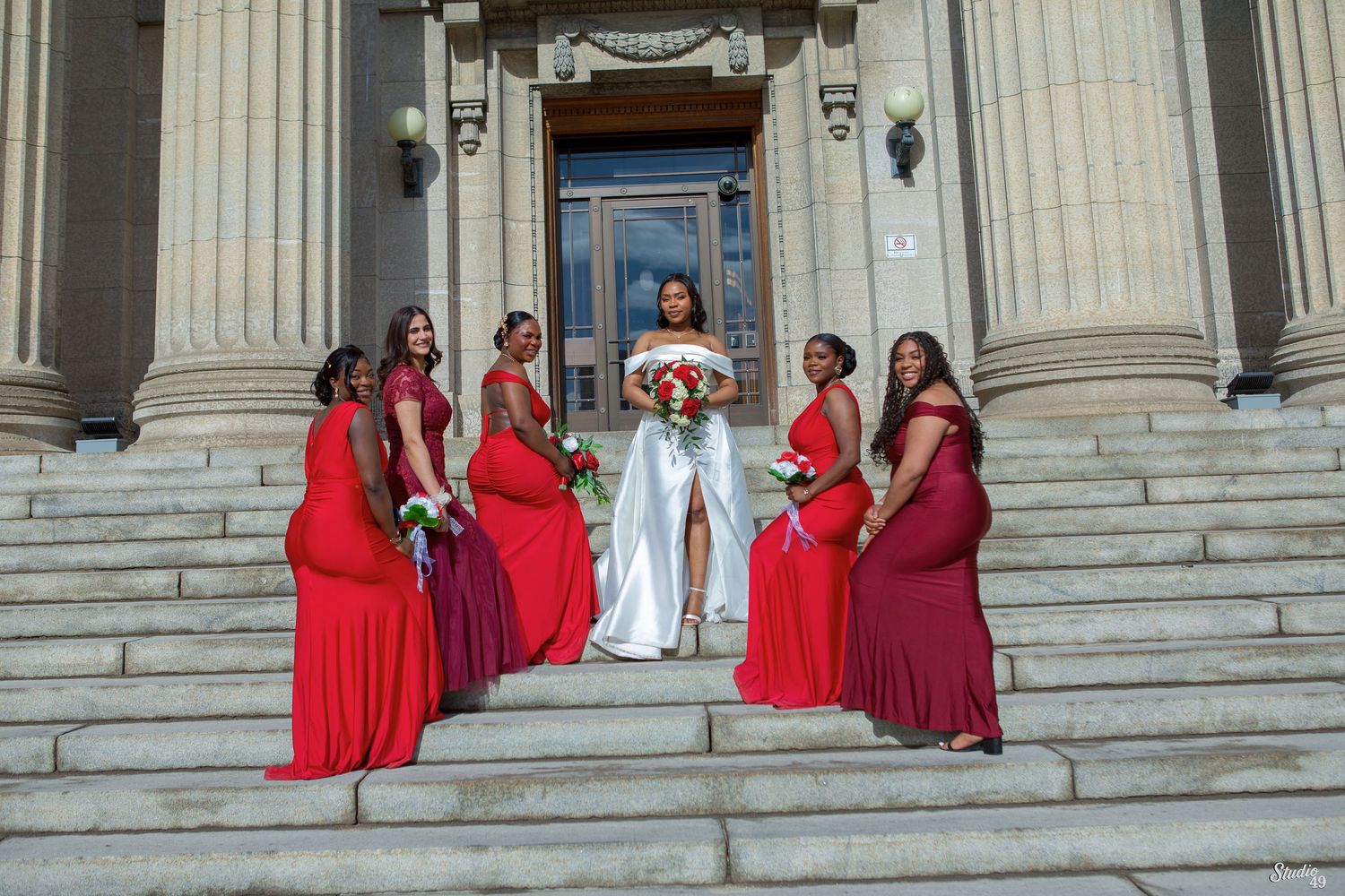 Bride in a white gown holding a bouquet poses on stone steps with five bridesmaids in red dresses outside a columned building.