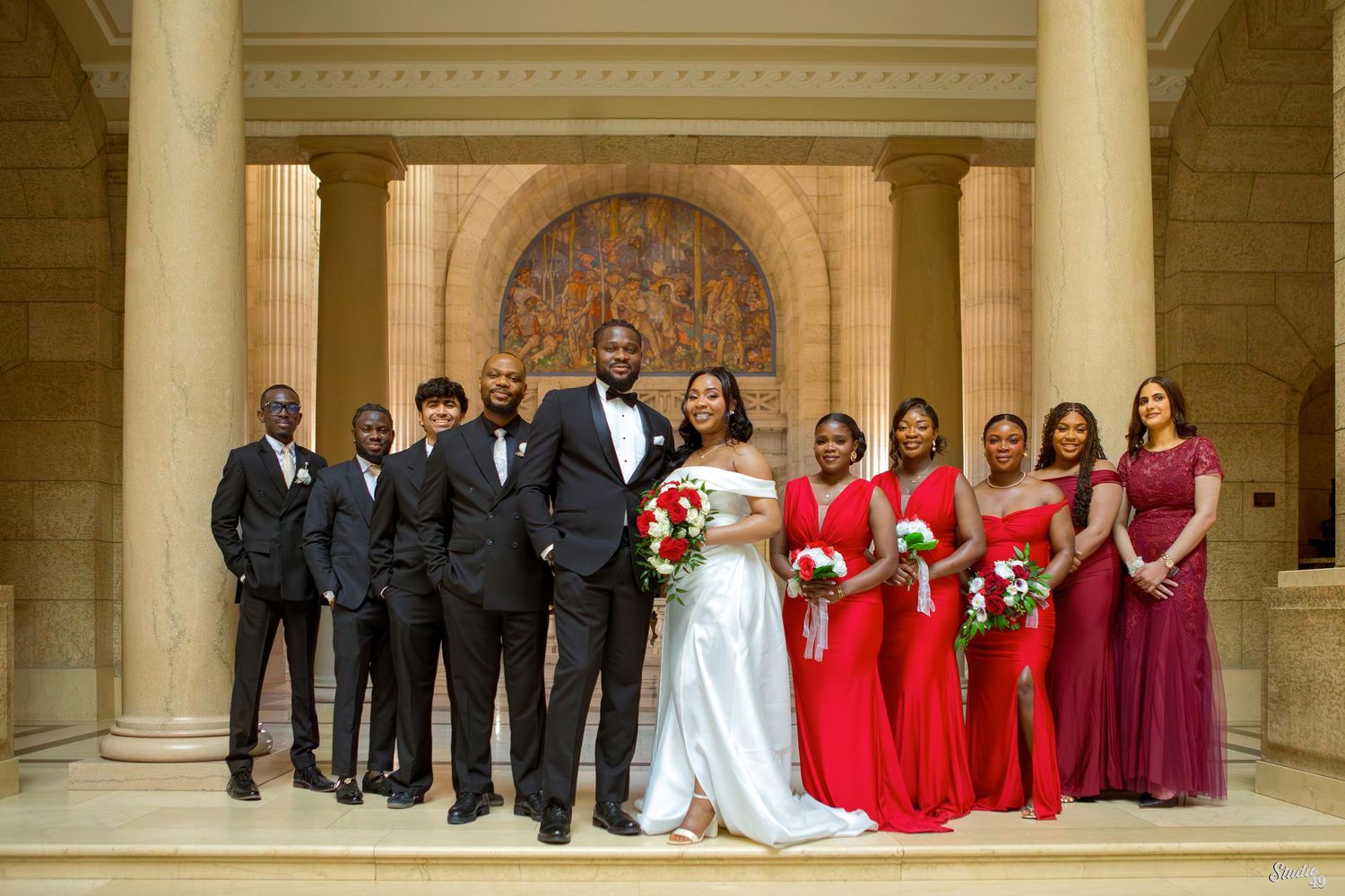 Bride in a white gown with a bouquet stands between groomsmen in dark suits and bridesmaids in red dresses in a grand stone hall.