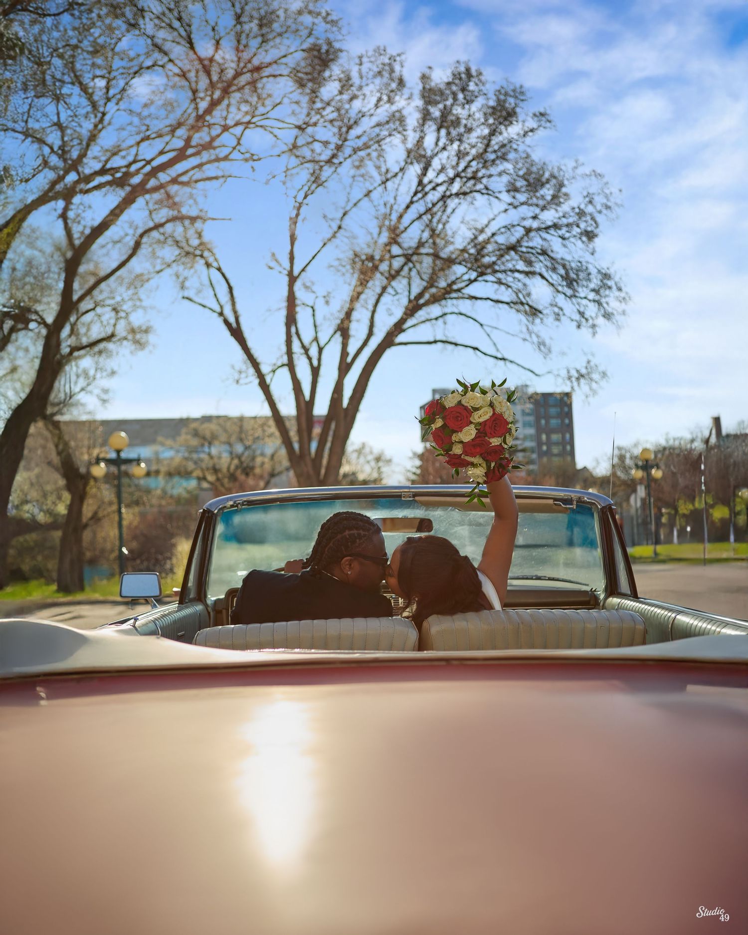 Newlywed couple kissing in a vintage convertible while the bride raises a red and white bouquet under bare trees and a blue sky.