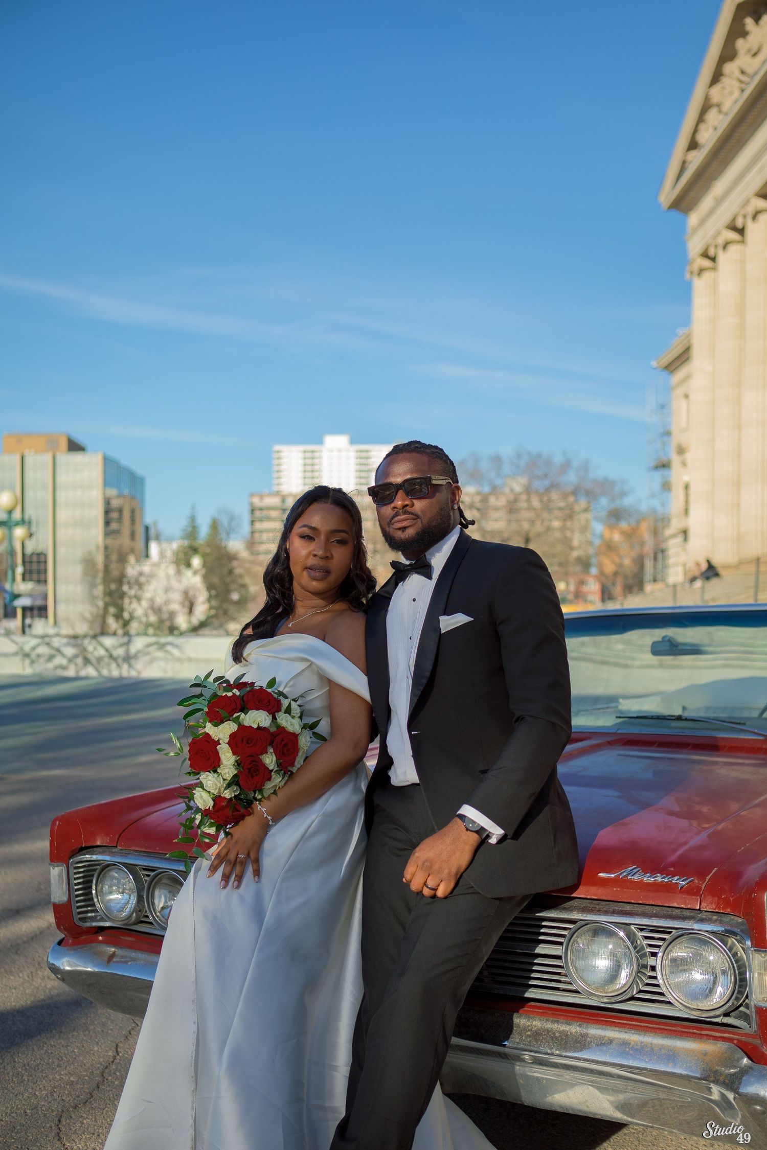 Bride in a white off-shoulder gown holding red roses poses beside a groom in a black tuxedo and sunglasses leaning on a vintage red car.