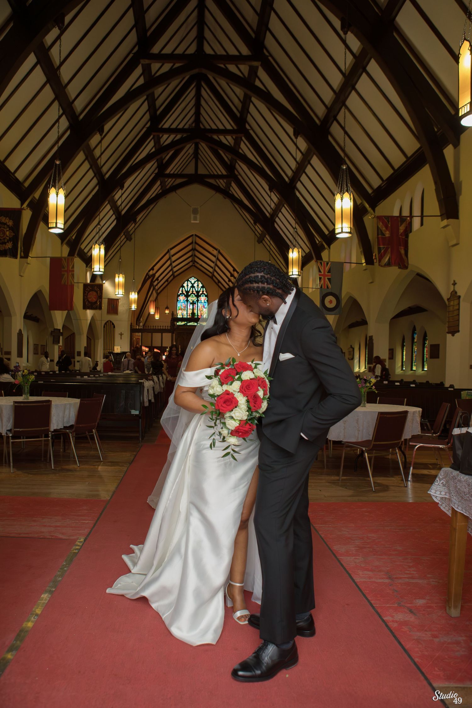 Bride and groom share a kiss in a church aisle, she holding a red rose bouquet, beneath wooden beams and hanging lanterns.