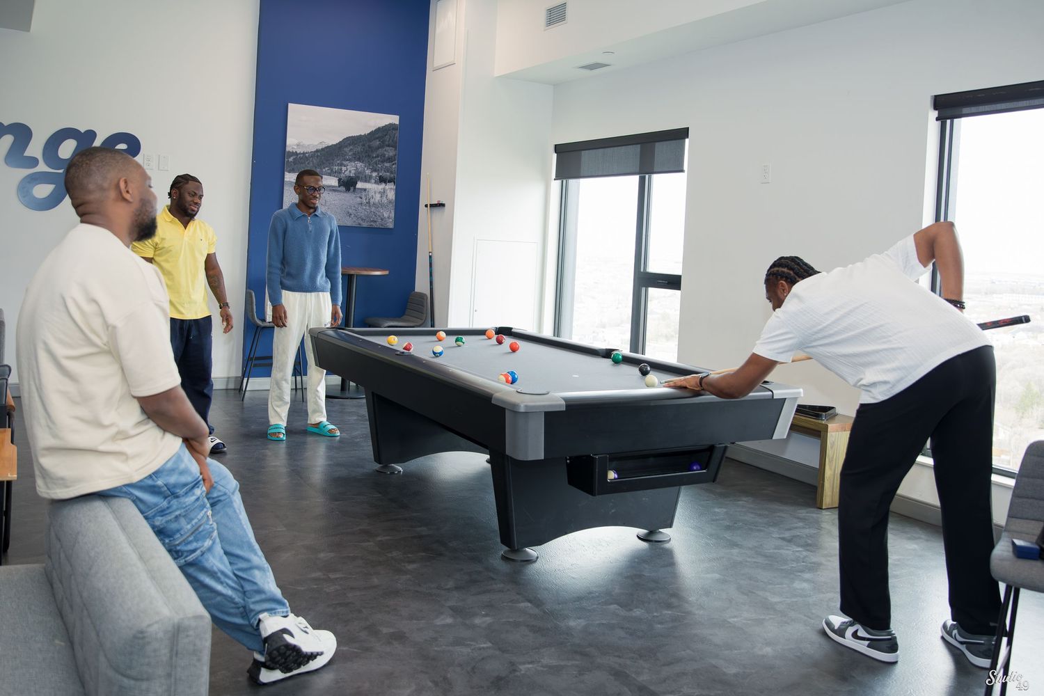 Four men gather in a bright modern lounge as one leans over a pool table to take a shot while the others watch.