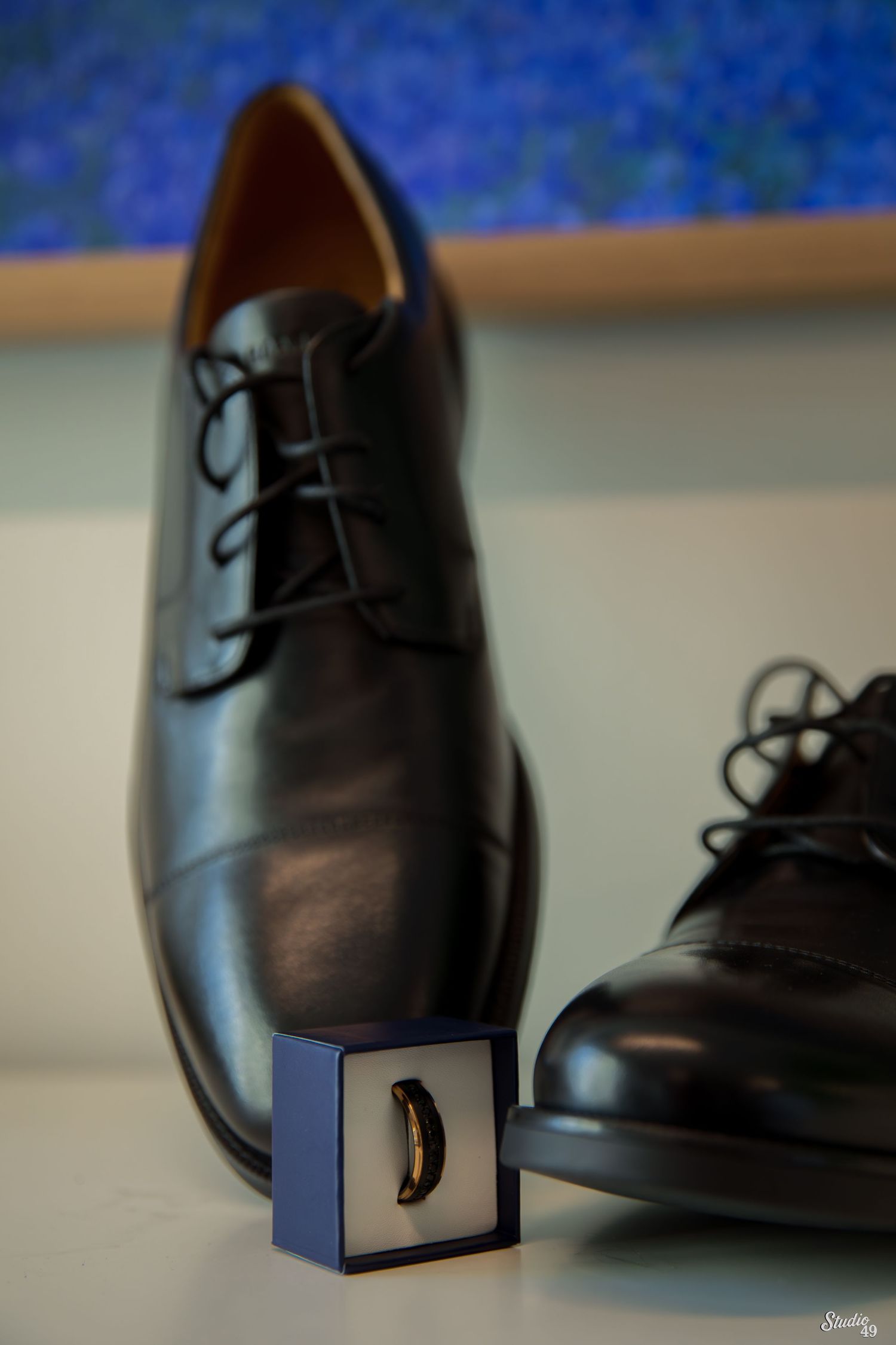 Polished black leather lace-up dress shoes beside a small navy ring box holding a men's wedding band on a white surface.