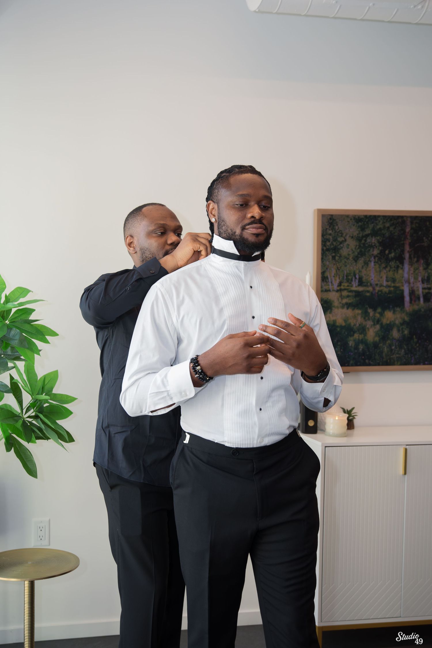 Groomsman helps a man in a white dress shirt and black trousers fasten his bow tie while getting ready for a wedding in a bright room.