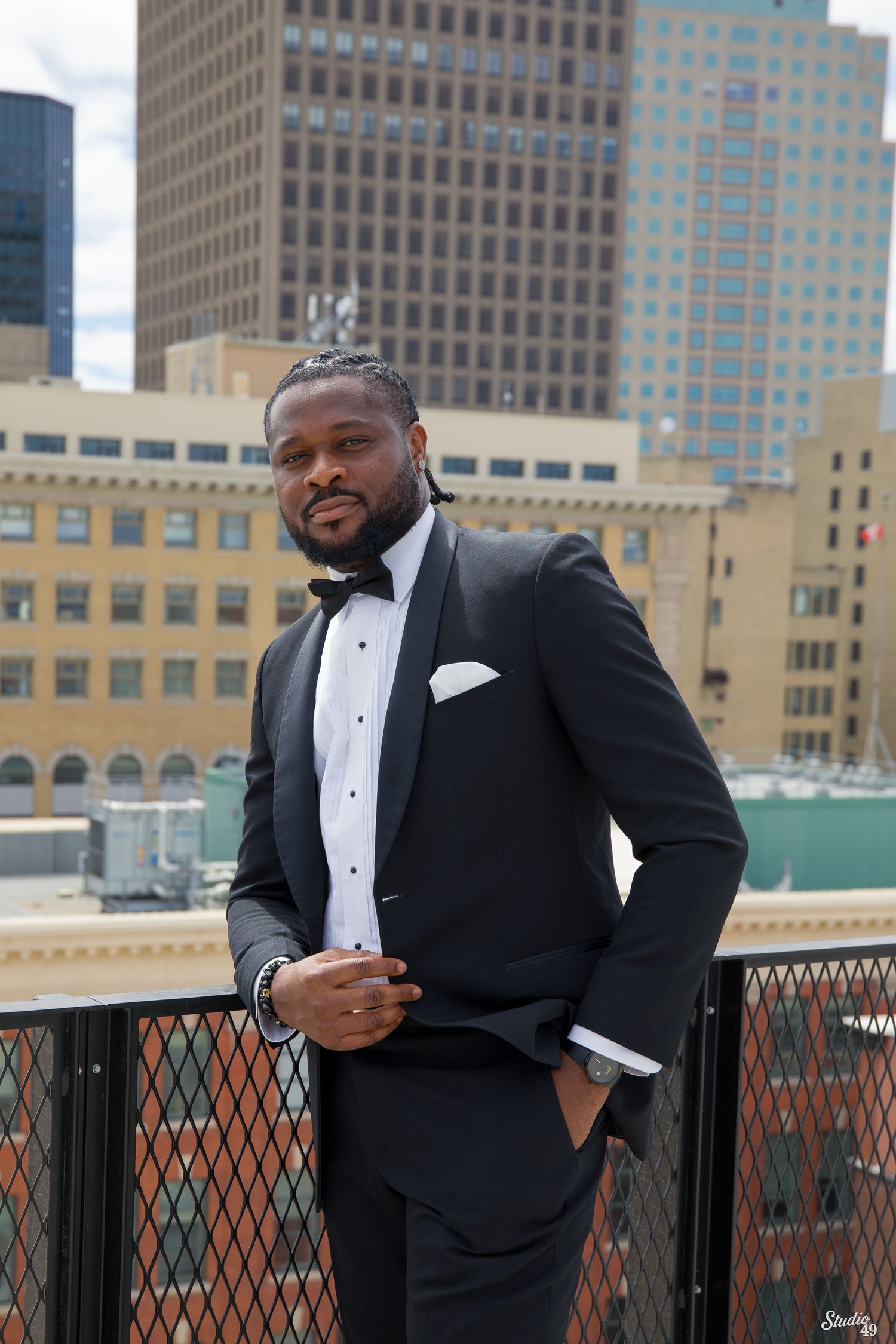 Bearded man in a black tuxedo with bow tie and white pocket square poses on a rooftop terrace with city skyscrapers behind him.
