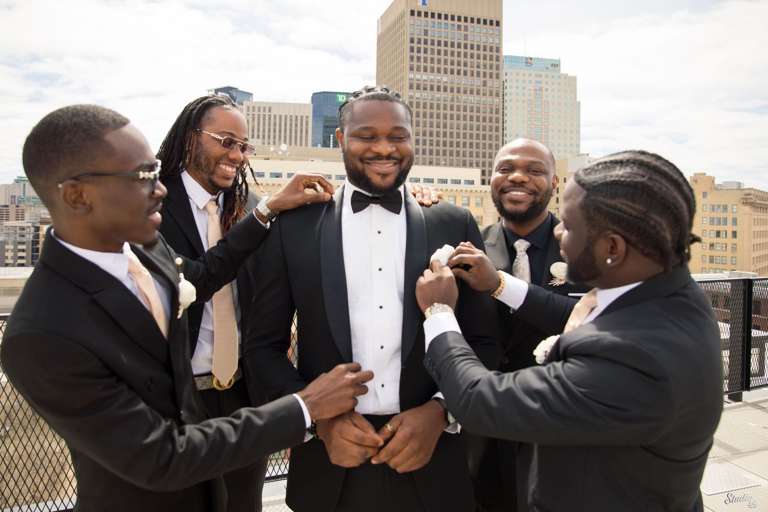 Groomsmen in black suits help a groom in a bow tie adjust his boutonniere and cuffs on a sunny rooftop with a city skyline behind them.