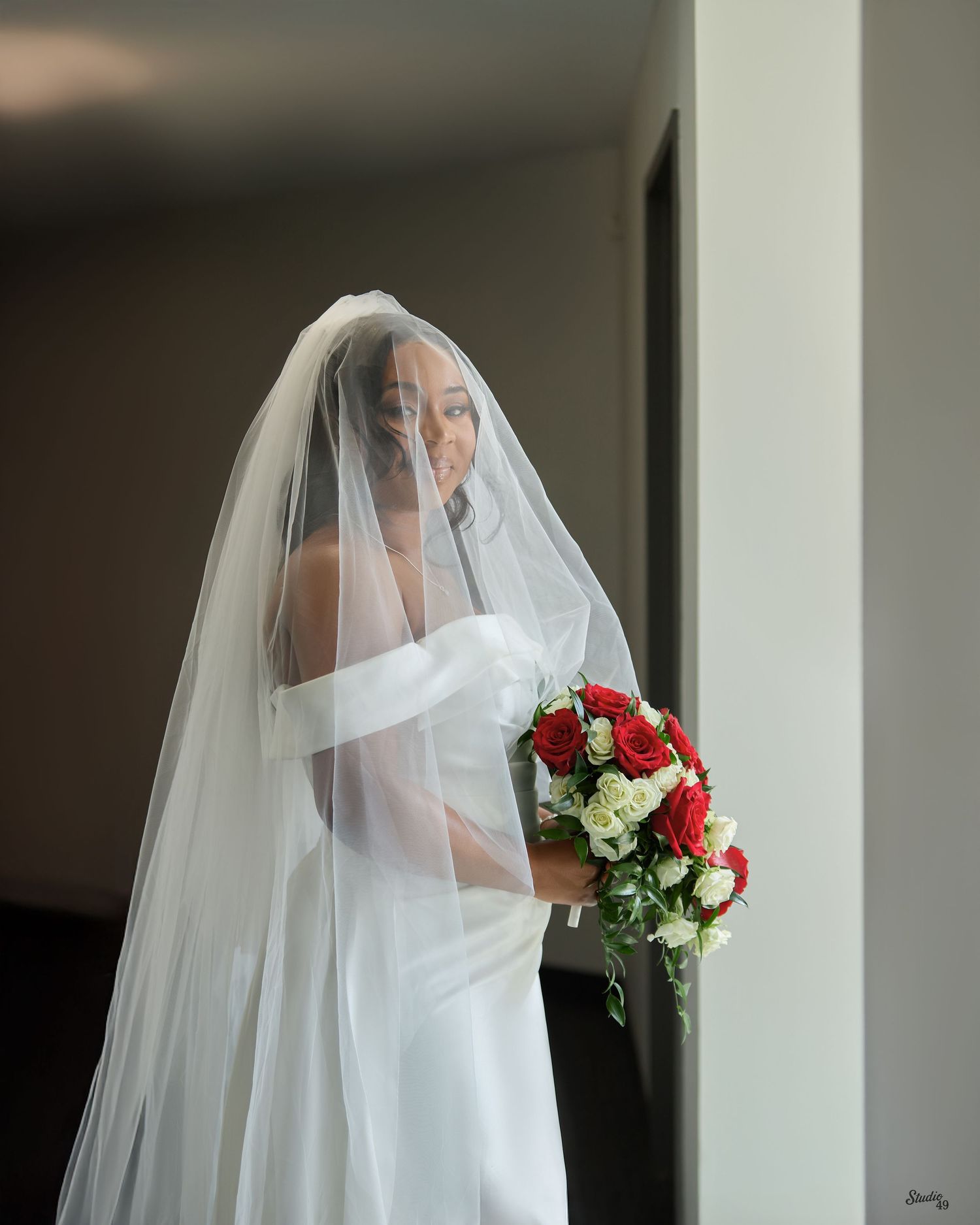 Bride in an elegant white off-shoulder gown and sheer veil holding a bouquet of red roses and white flowers beside a window.
