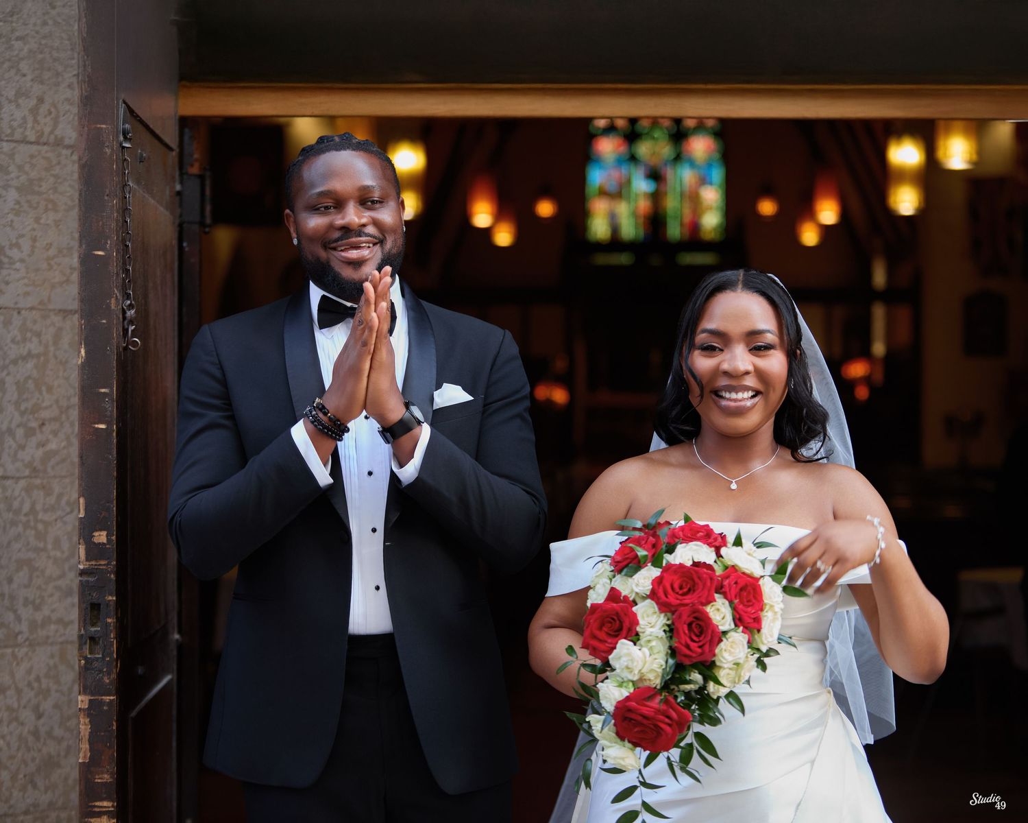 Smiling groom in a black tuxedo and bow tie with hands pressed together beside a joyful bride in a white gown holding a red rose bouquet.