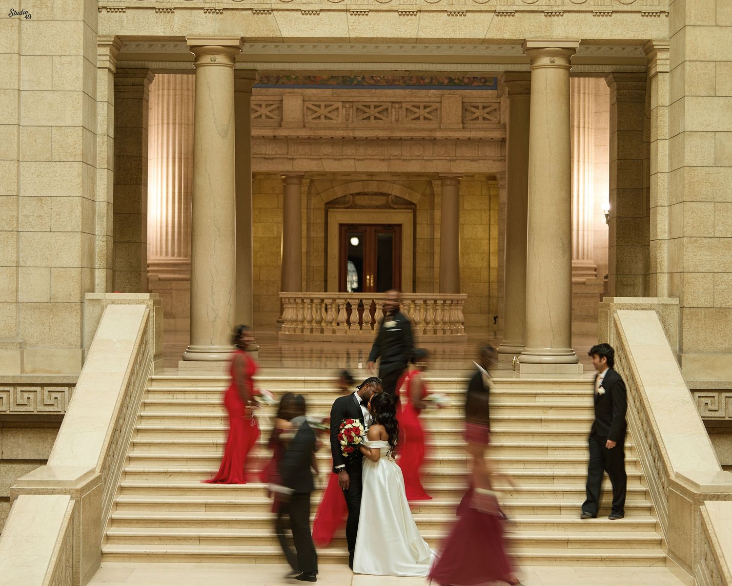 Wedding party in red dresses and black suits poses with a bride in white on grand stone steps of a columned building.