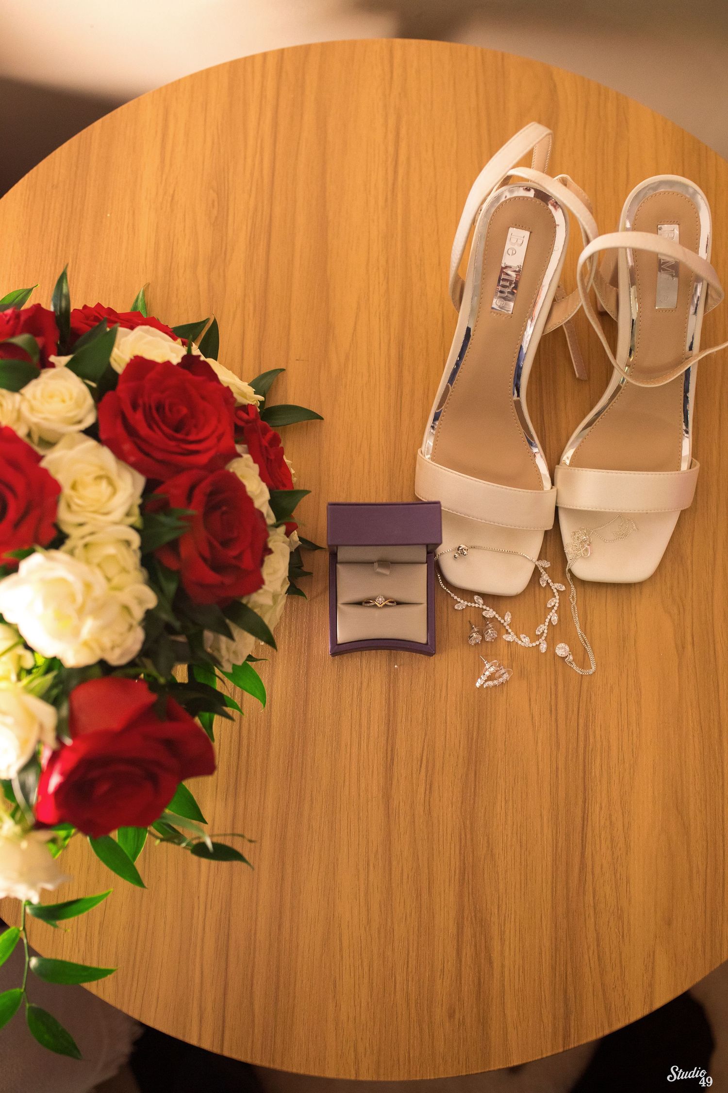 Red and white rose bridal bouquet beside a ring box, pearl necklace, and nude strappy heels on a round wooden table.