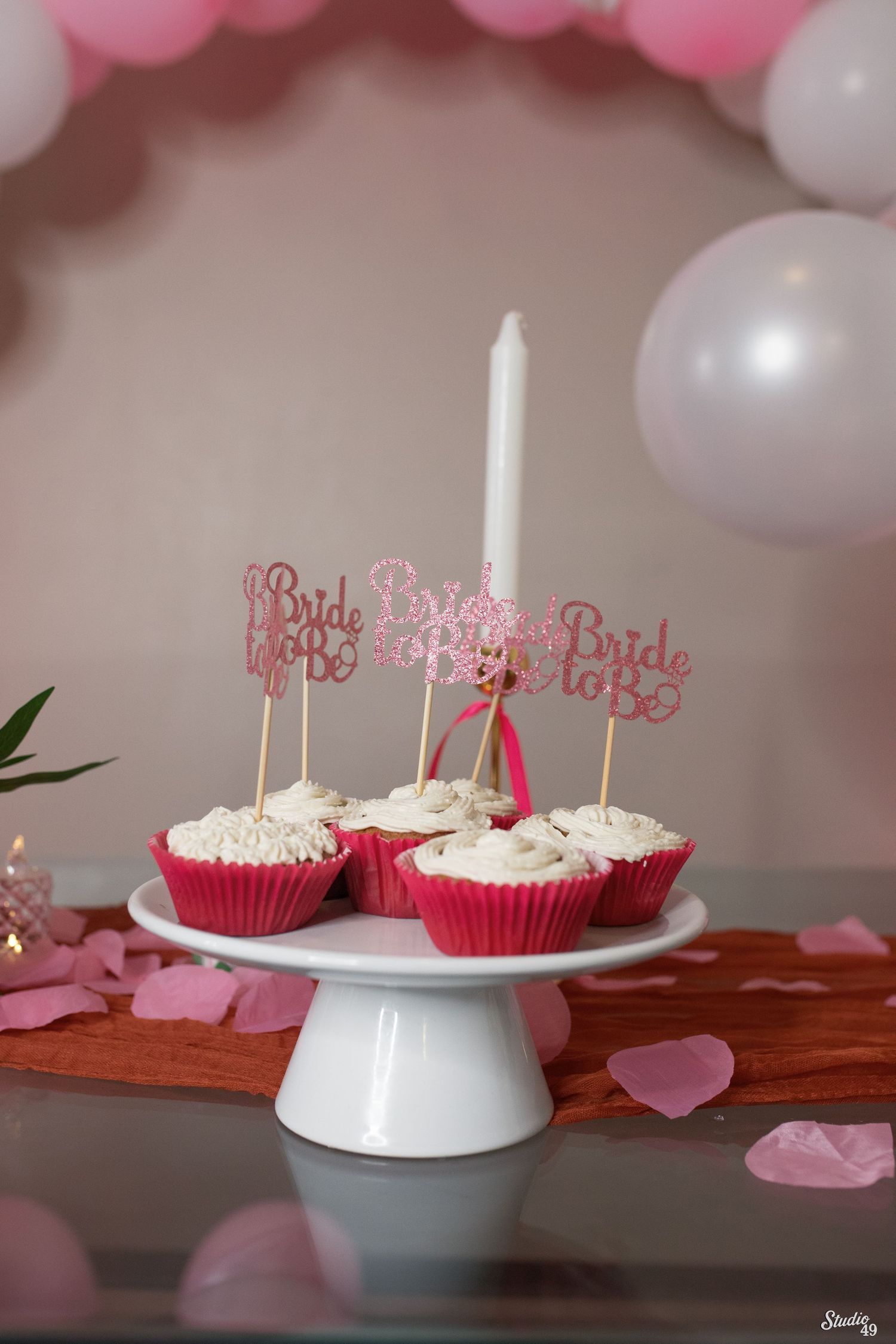 Pink bride to be cupcakes with frosting and glitter toppers on a white cake stand, surrounded by rose petals and balloons.