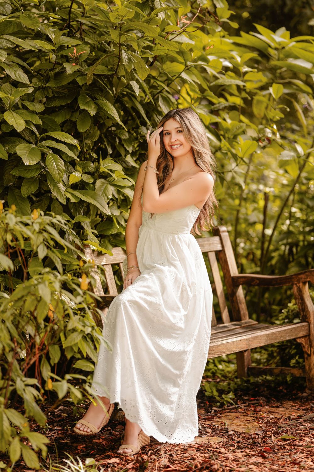 Young woman in white sundress sitting on wooden bench surrounded by lush green foliage in a garden setting.