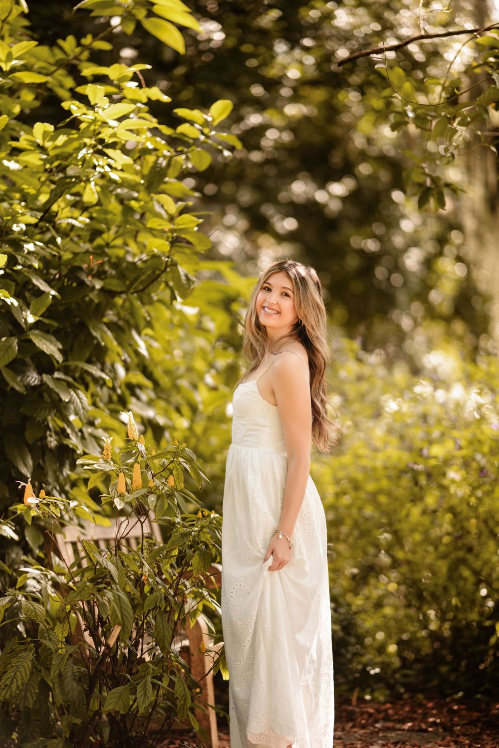 Young woman in white dress smiling outdoors among lush green trees in golden sunlight.