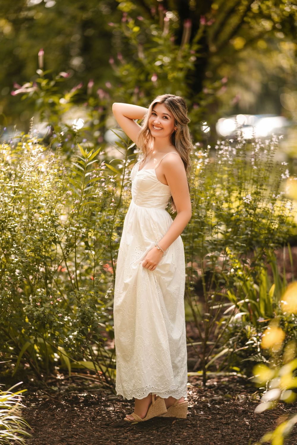 Young woman in white lace maxi dress posing gracefully among lush green garden plants in warm sunlight.