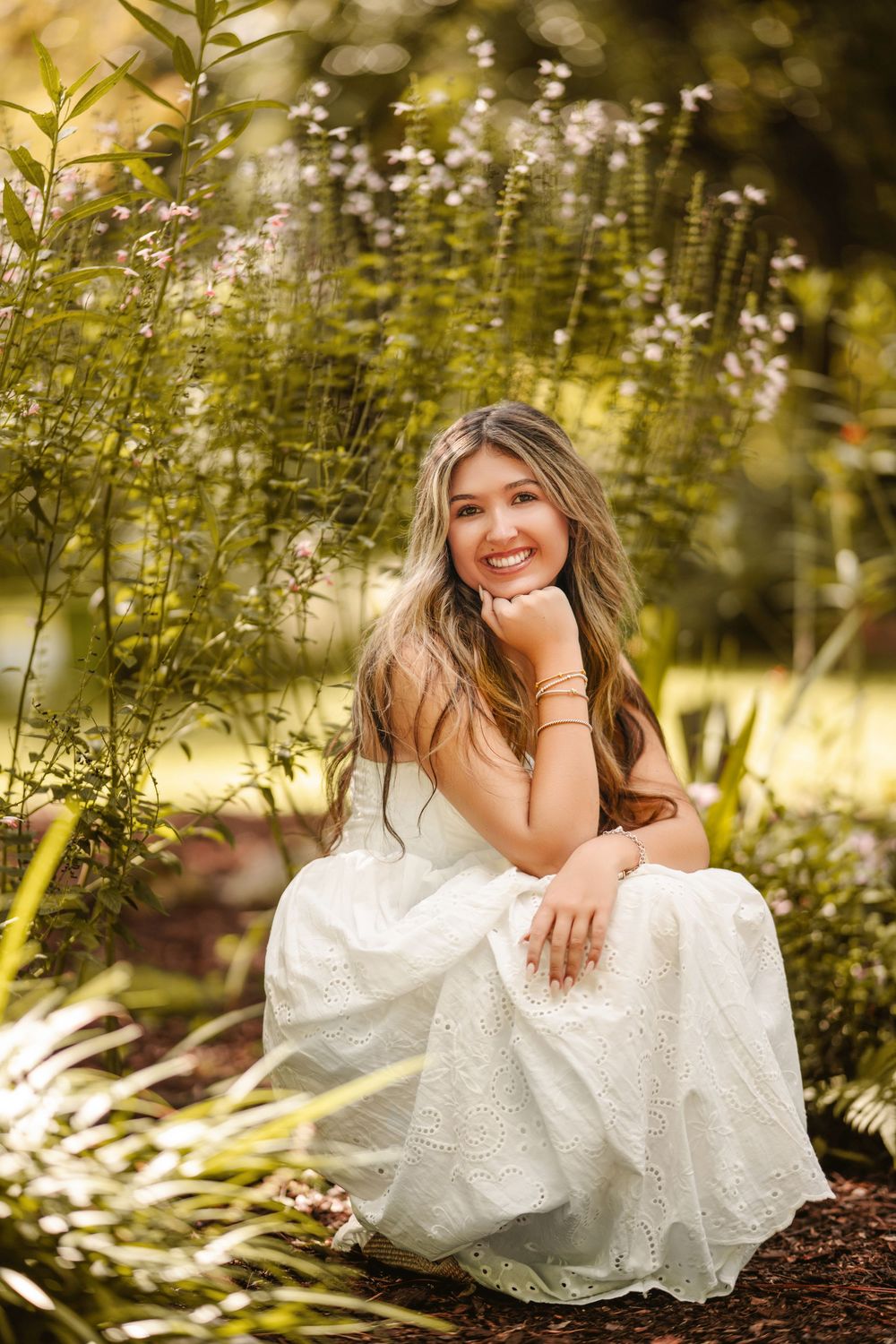Young woman in white dress sitting among wildflowers in a sunlit garden, smiling warmly at the camera.