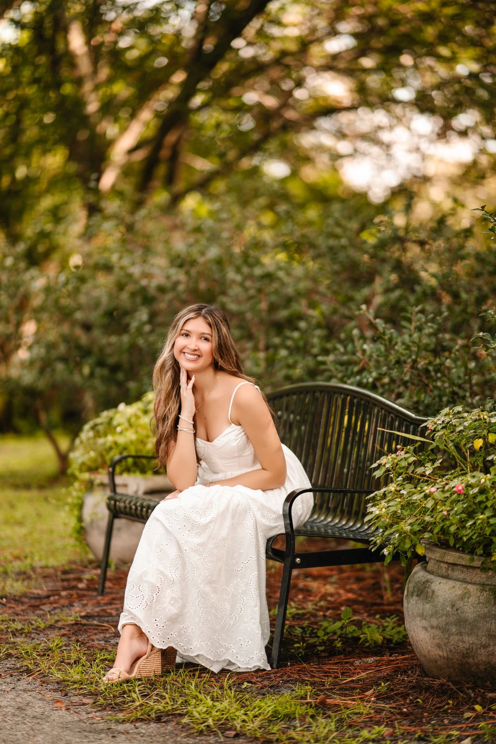 Young woman in white dress sitting gracefully on a garden bench, smiling warmly, surrounded by lush green trees and plants.