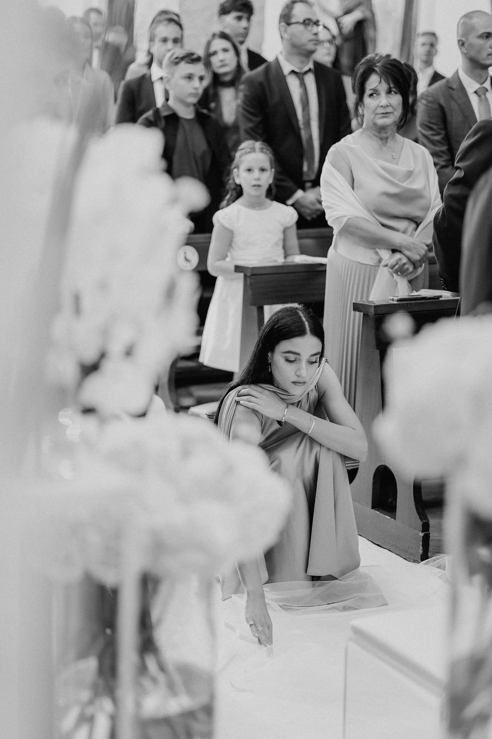 Gli invitati e una bambina seguono la cerimonia in chiesa, in bianco e nero.