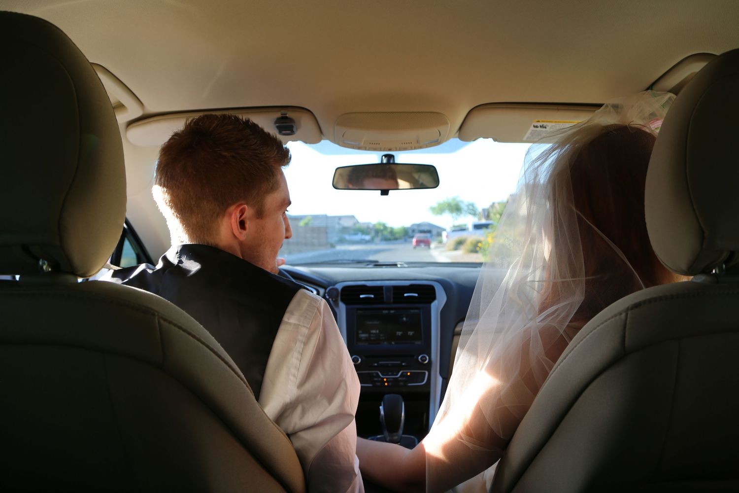 A groom in a vest and white shirt sits in the driver's seat while a bride in a long veil rides beside him in a car.
