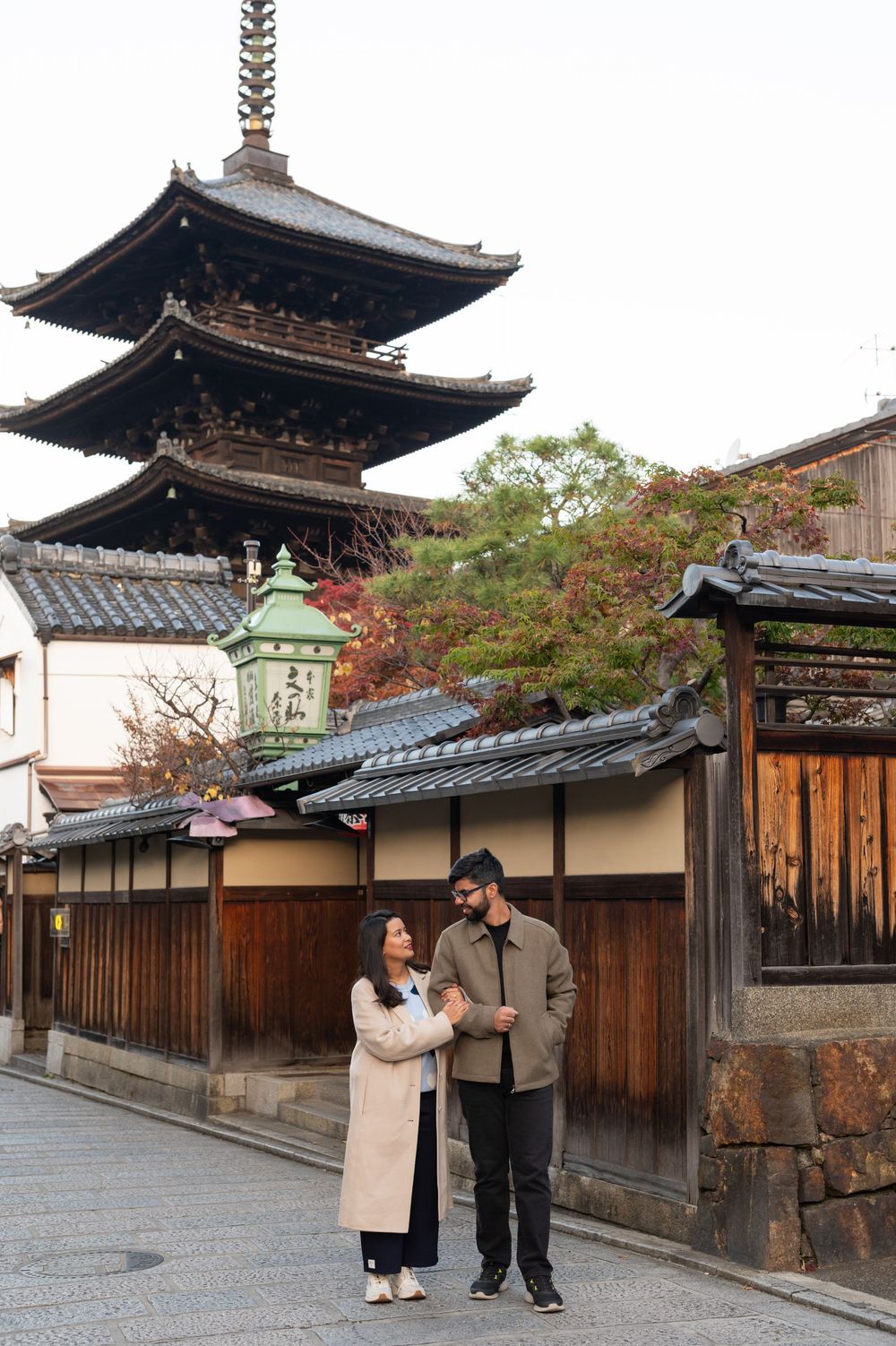 Couple in autumn coats walk together on a quiet Kyoto street beside wooden fences with the Yasaka Pagoda rising in the background.