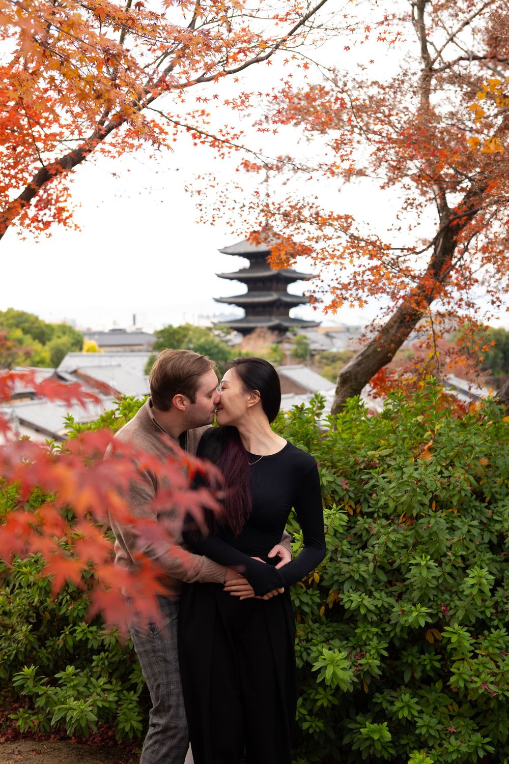 Couple kissing among vibrant red autumn maple leaves with a traditional Japanese pagoda rising in the background.