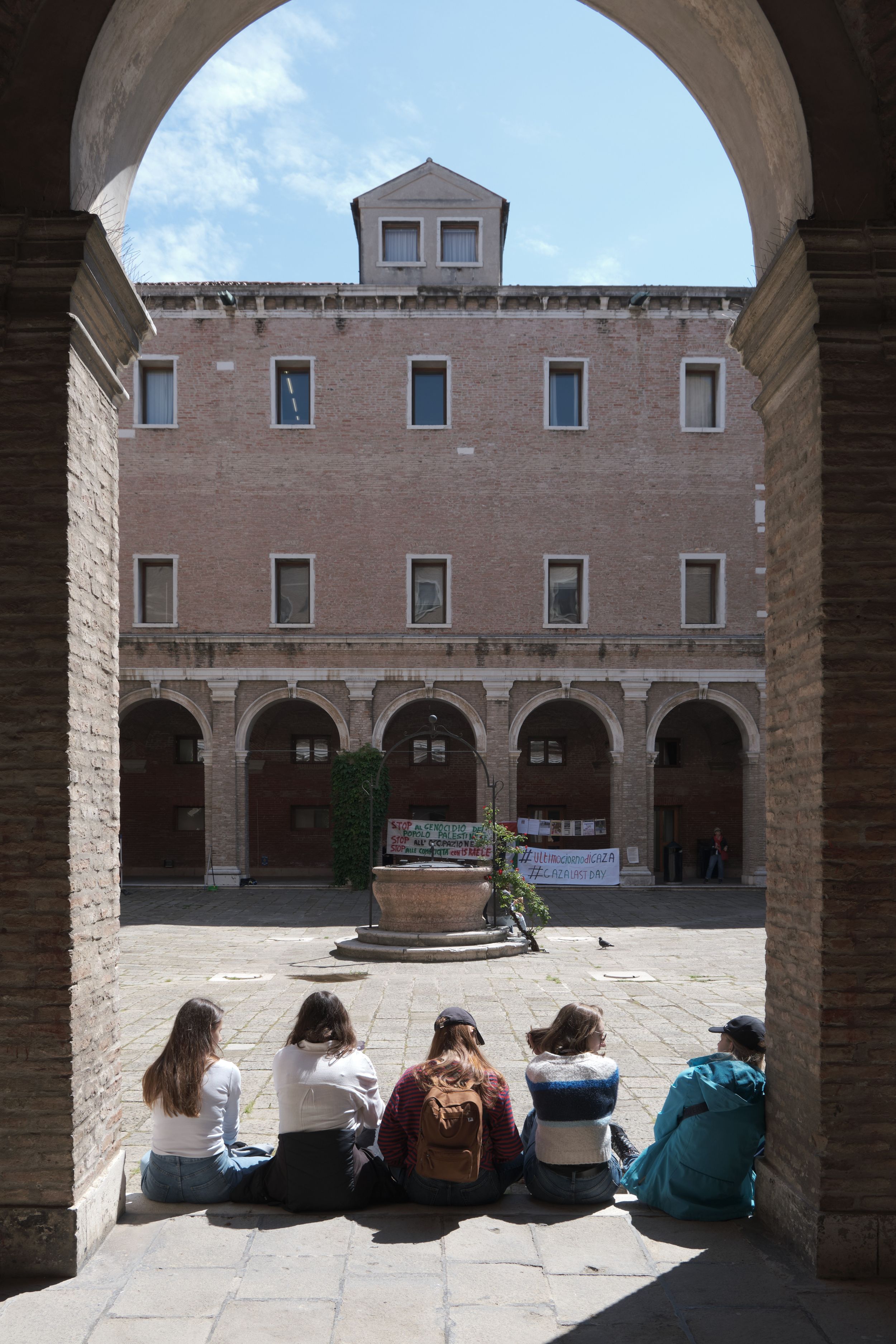 Five students sit on the stone ground in a sunlit brick courtyard, framed by an archway, with a fountain and arcaded building ahead.