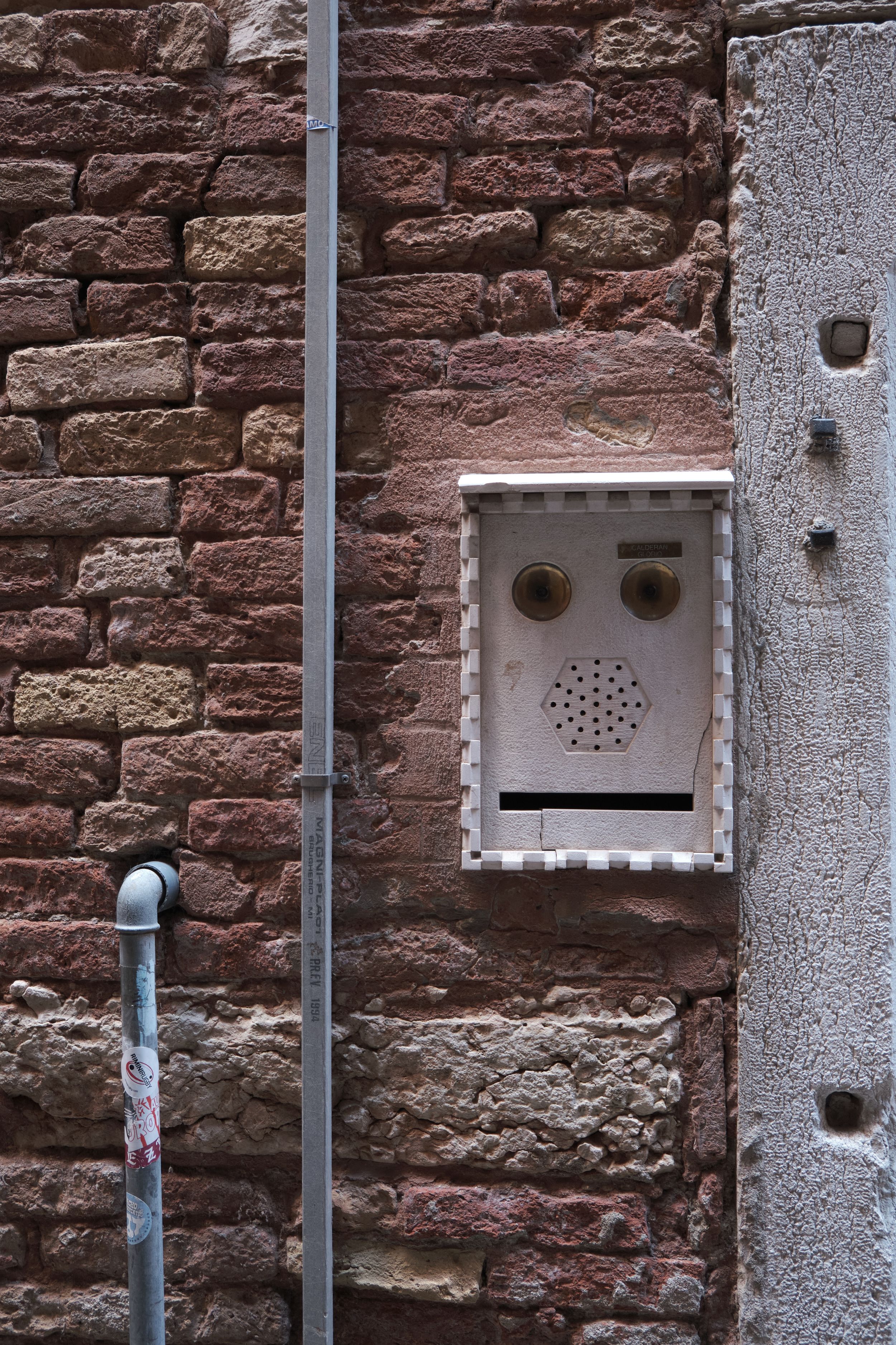 Weathered intercom panel with speaker grille and two buttons mounted on an old red brick wall beside a metal drainpipe.