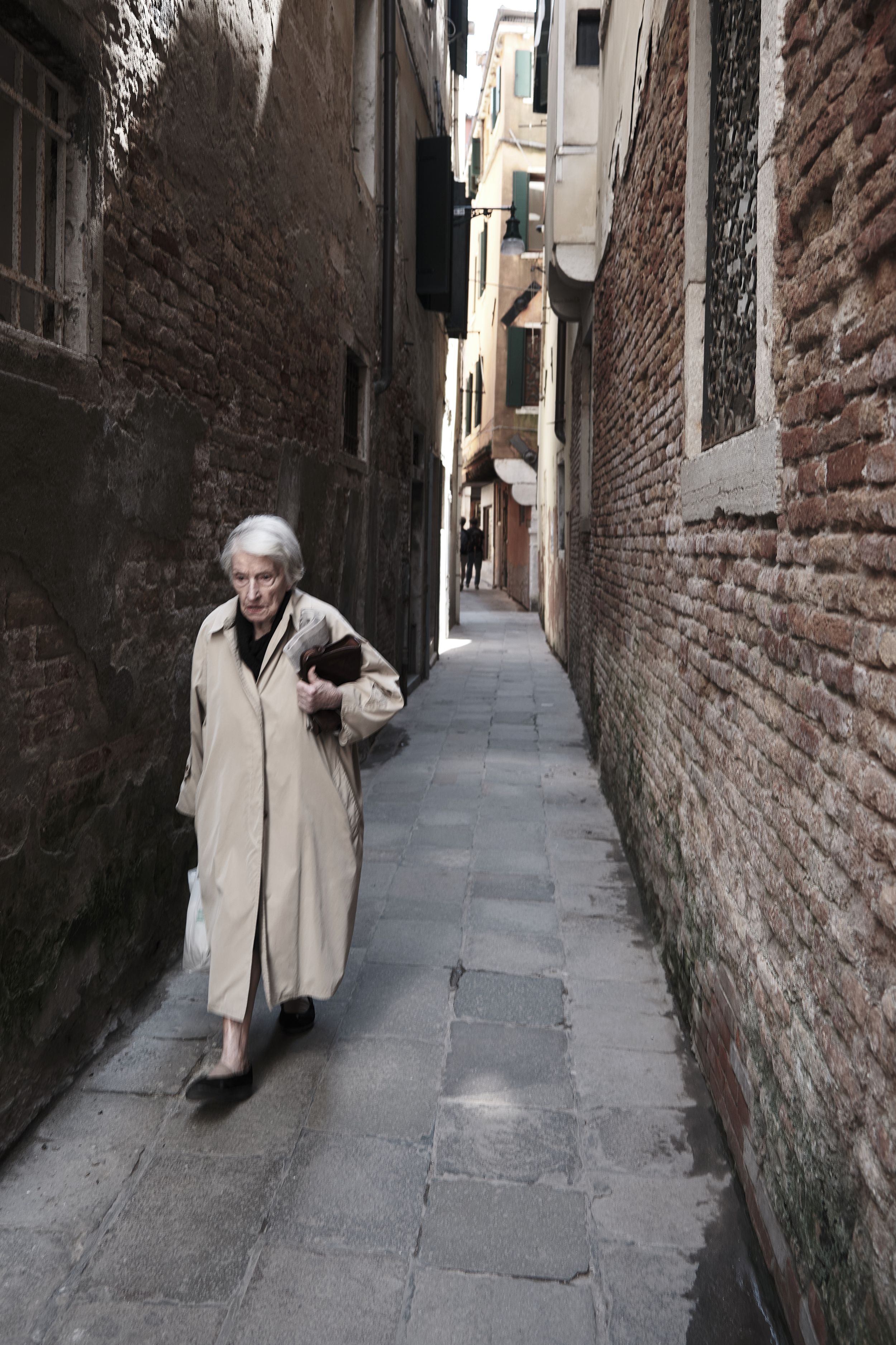 Elderly woman in a beige trench coat walks down a narrow brick alley in Venice, Italy, carrying a book.
