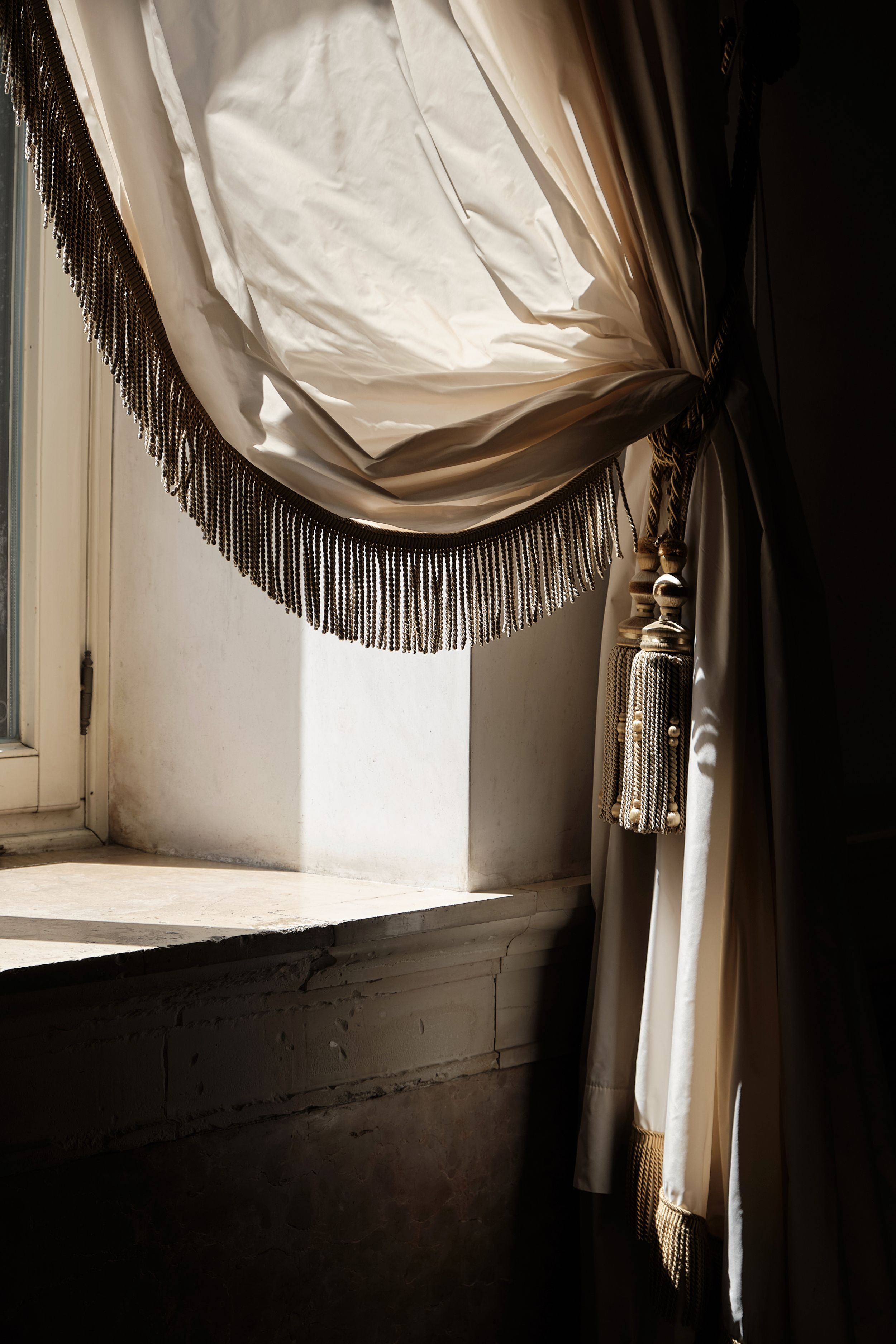 Sunlight streams through a window, illuminating an elegant cream silk curtain with beaded fringe trim and a decorative tassel tieback.