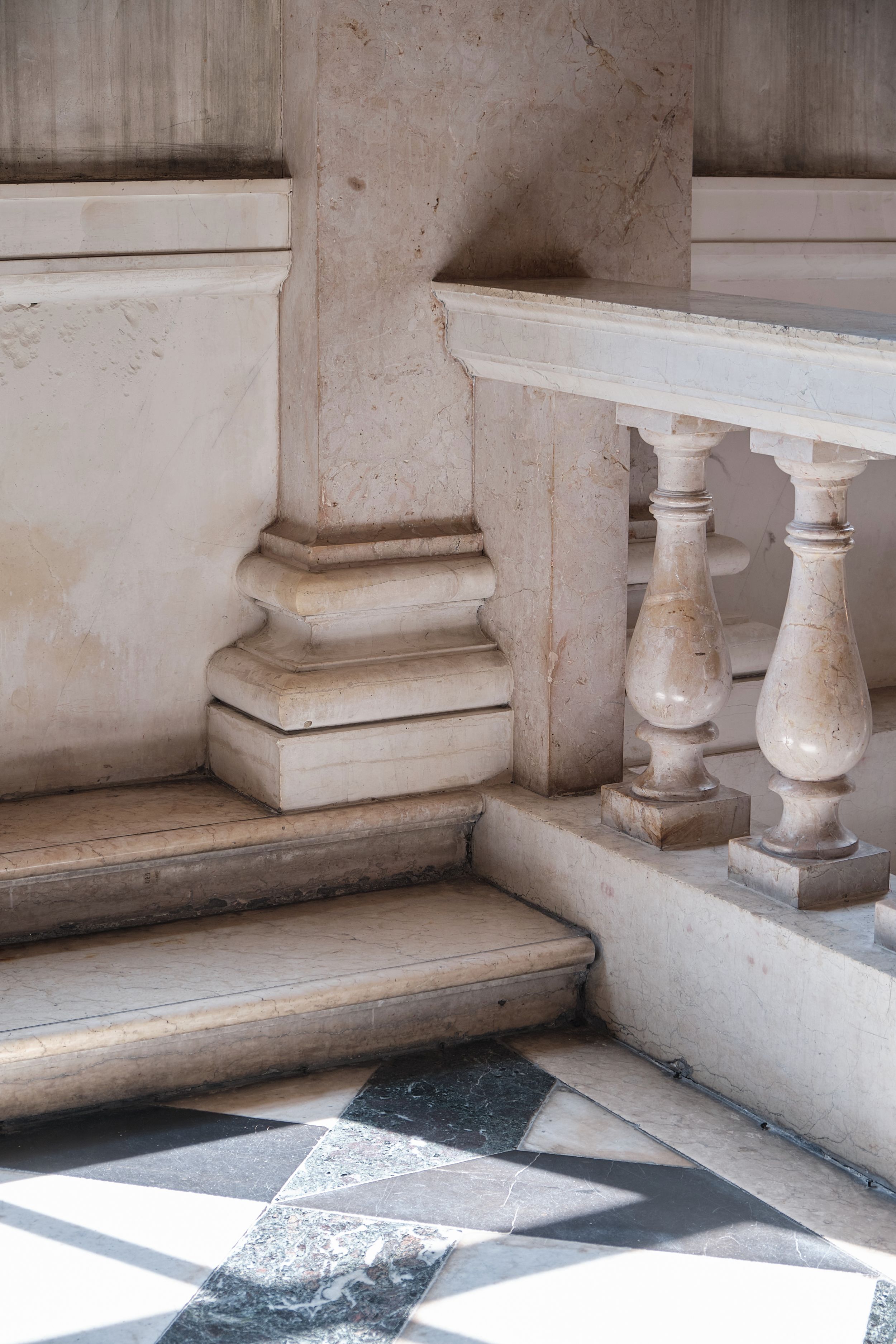 Marble staircase with carved balusters and a pilaster base above black and white checkered stone flooring in a classical interior.