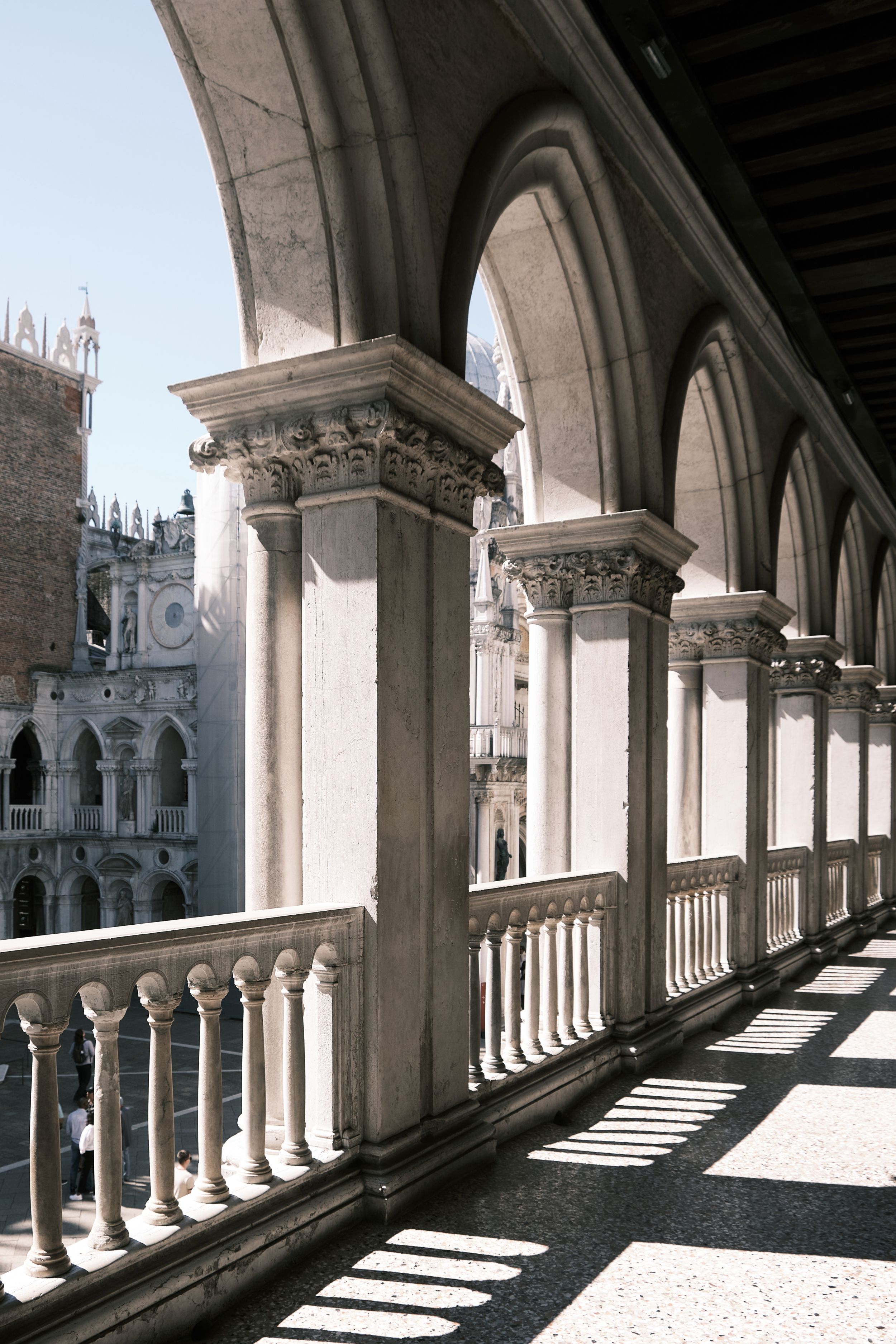 Sunlit Gothic arcade with pointed arches, carved column capitals and a stone balustrade overlooking an ornate palace courtyard in Venice.