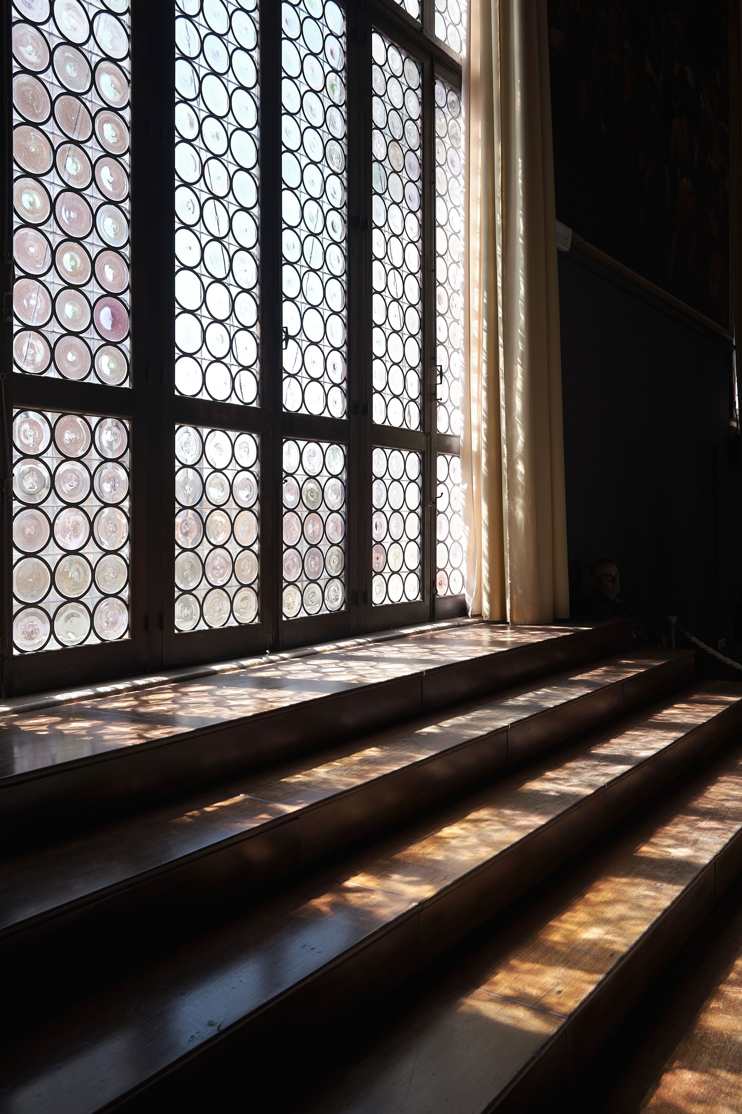 Sunlight streams through a leaded glass window with circular panes, casting warm patterns across polished wooden steps in a dim interior.