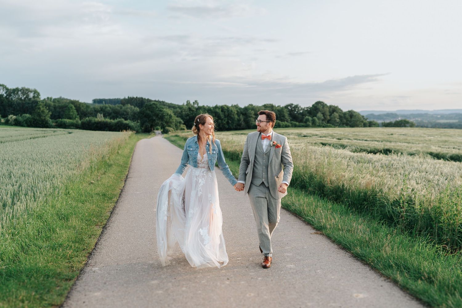 Bride and groom holding hands and walking down a country path between green fields on their wedding day, with trees in the distance.