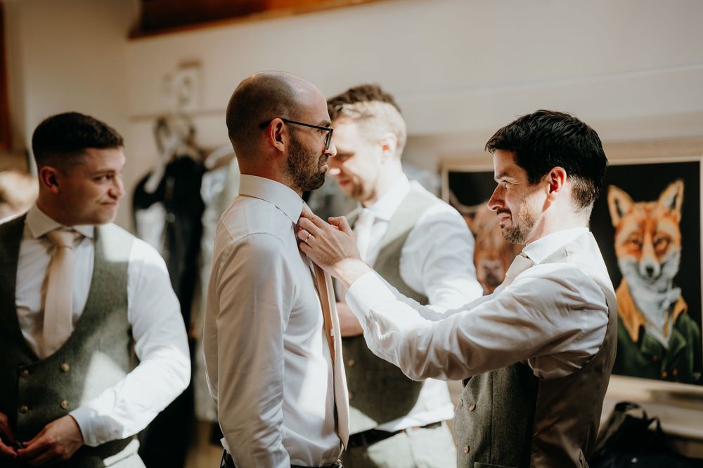 Groomsmen helping groom adjust his tie during wedding morning preparations in rustic venue.