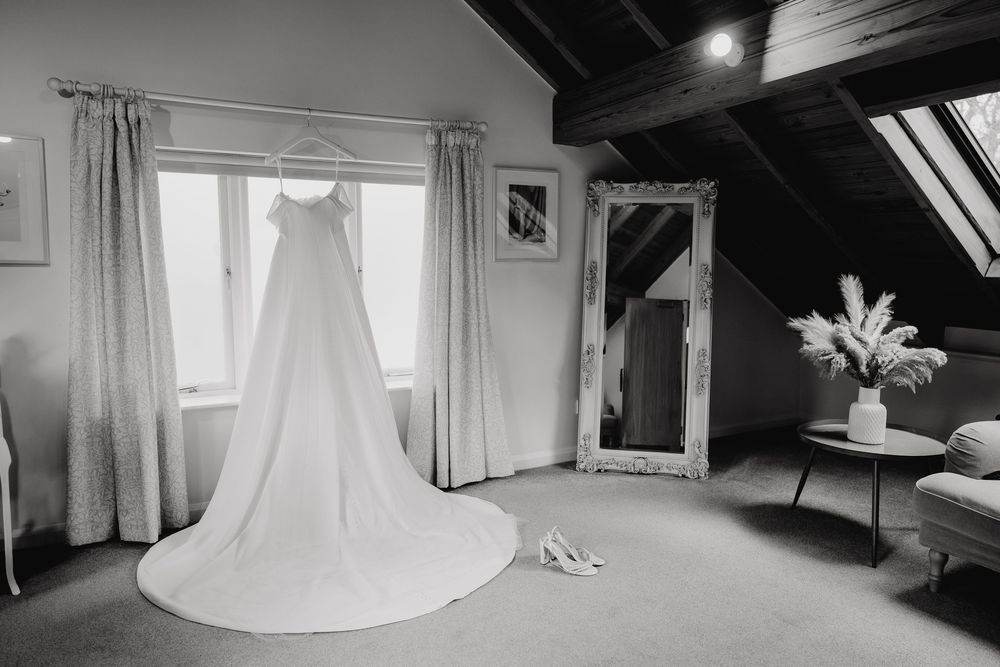 Black and white photo of a wedding dress hanging in an attic room with mirror and bridal shoes.
