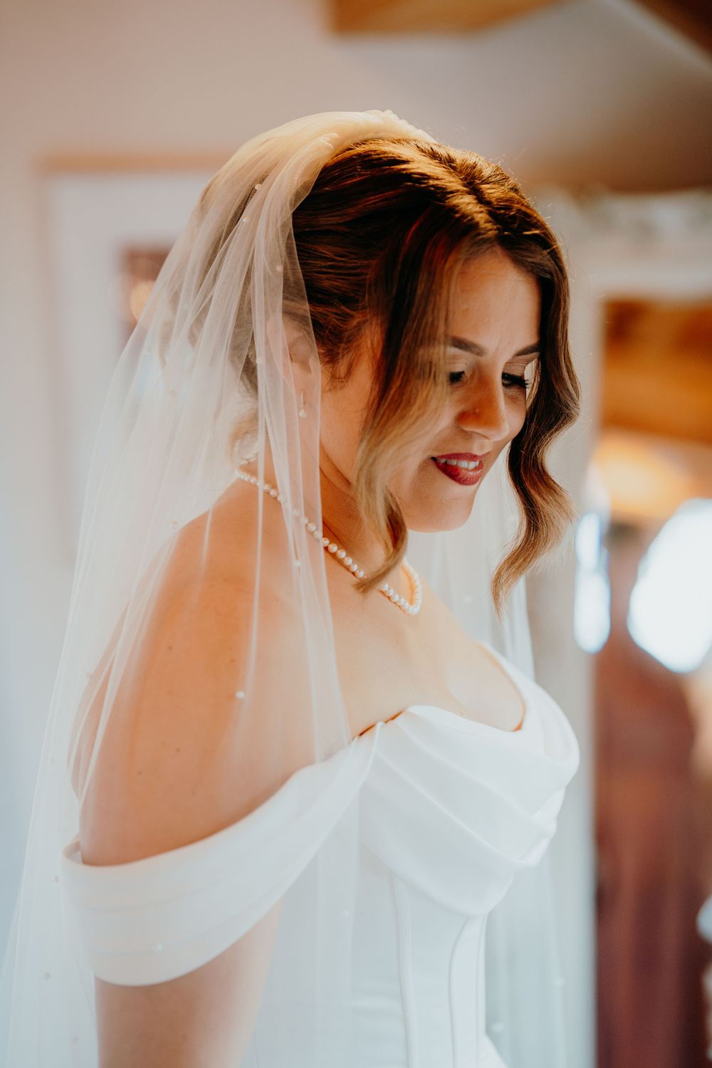 Bride in white off-shoulder gown and pearl necklace smiles softly while wearing a delicate veil.